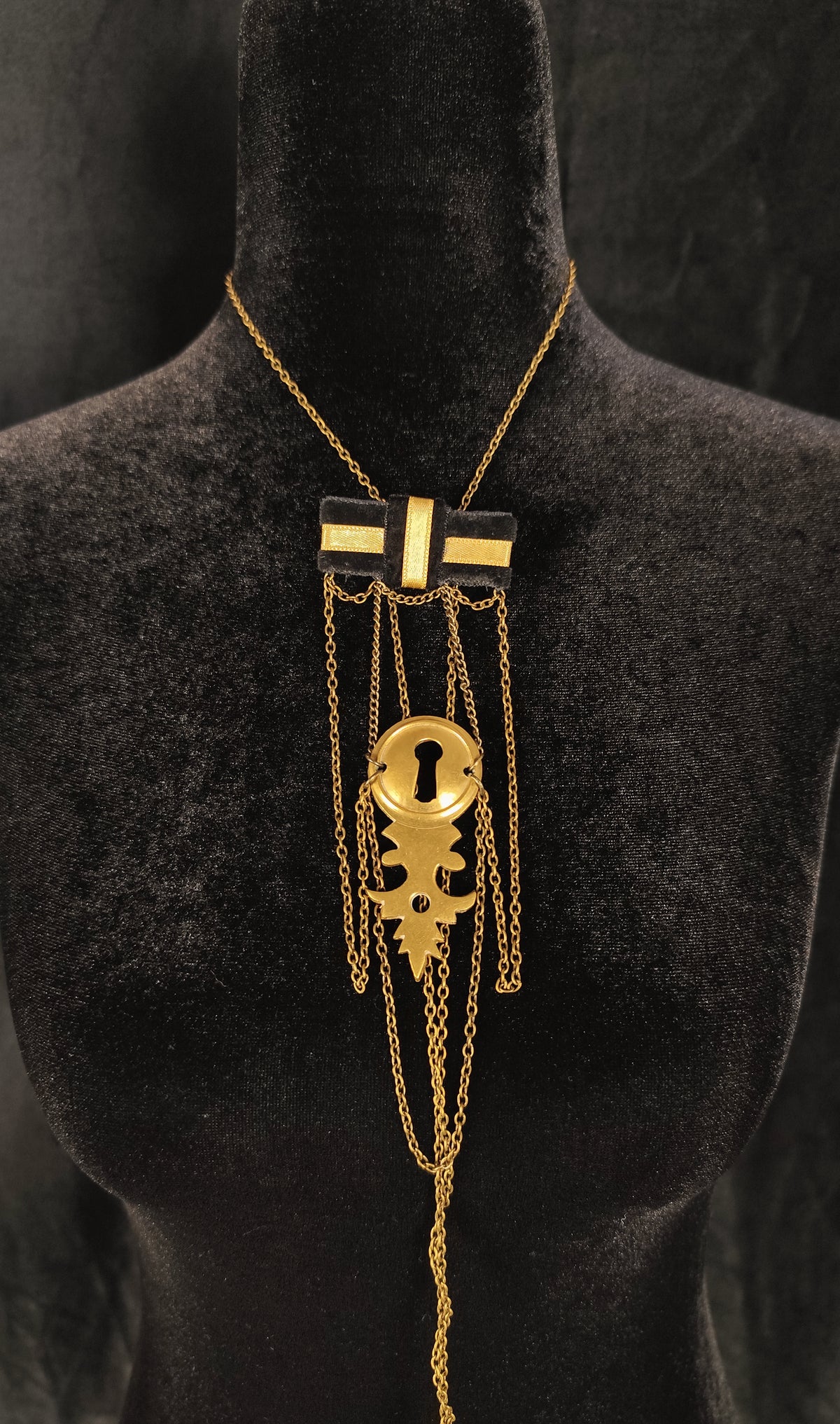 A gold and black statement necklace displayed on a black velvet mannequin bust. The necklace features a gold chain, a black and gold rectangular centerpiece, and a large gold keyhole pendant with dangling chains.