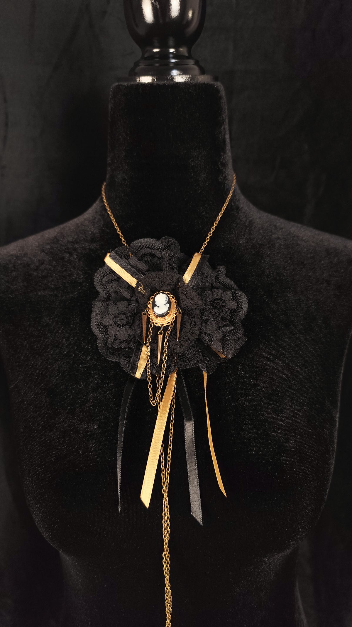 A gothic-style black lace brooch necklace with gold-tone chains, ribbons, and a central cameo, displayed on a black velvet mannequin bust.