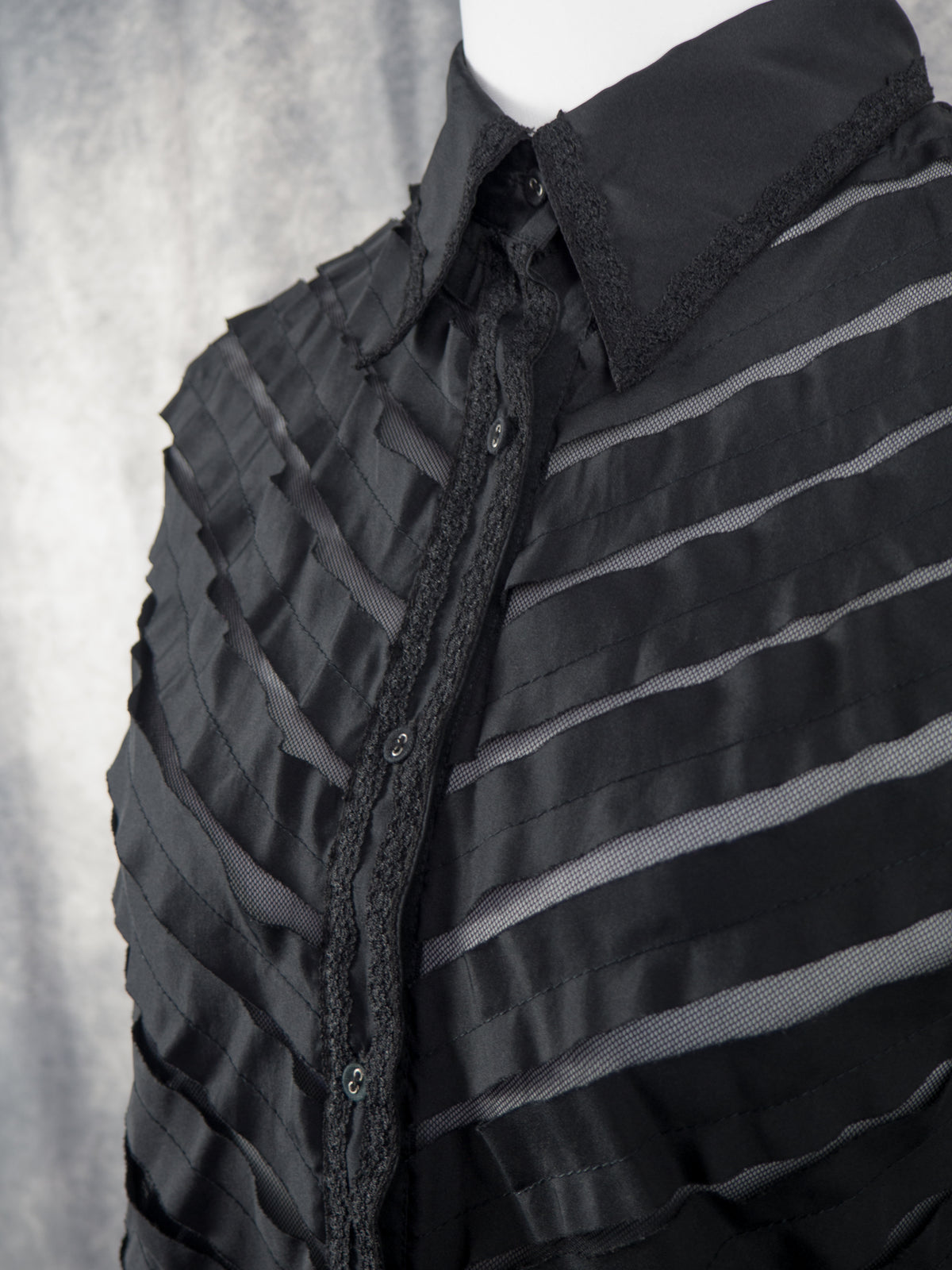 A close-up, angled view of the front upper section of the black blouse on the mannequin. The focus is on the collar, neckline, and button placket, which are all detailed with a delicate black lace trim along the edges. The semi-sheer bodice features alternating diagonal bands of solid fabric and sheer mesh, showing the texture and construction of the chevron pattern in detail.