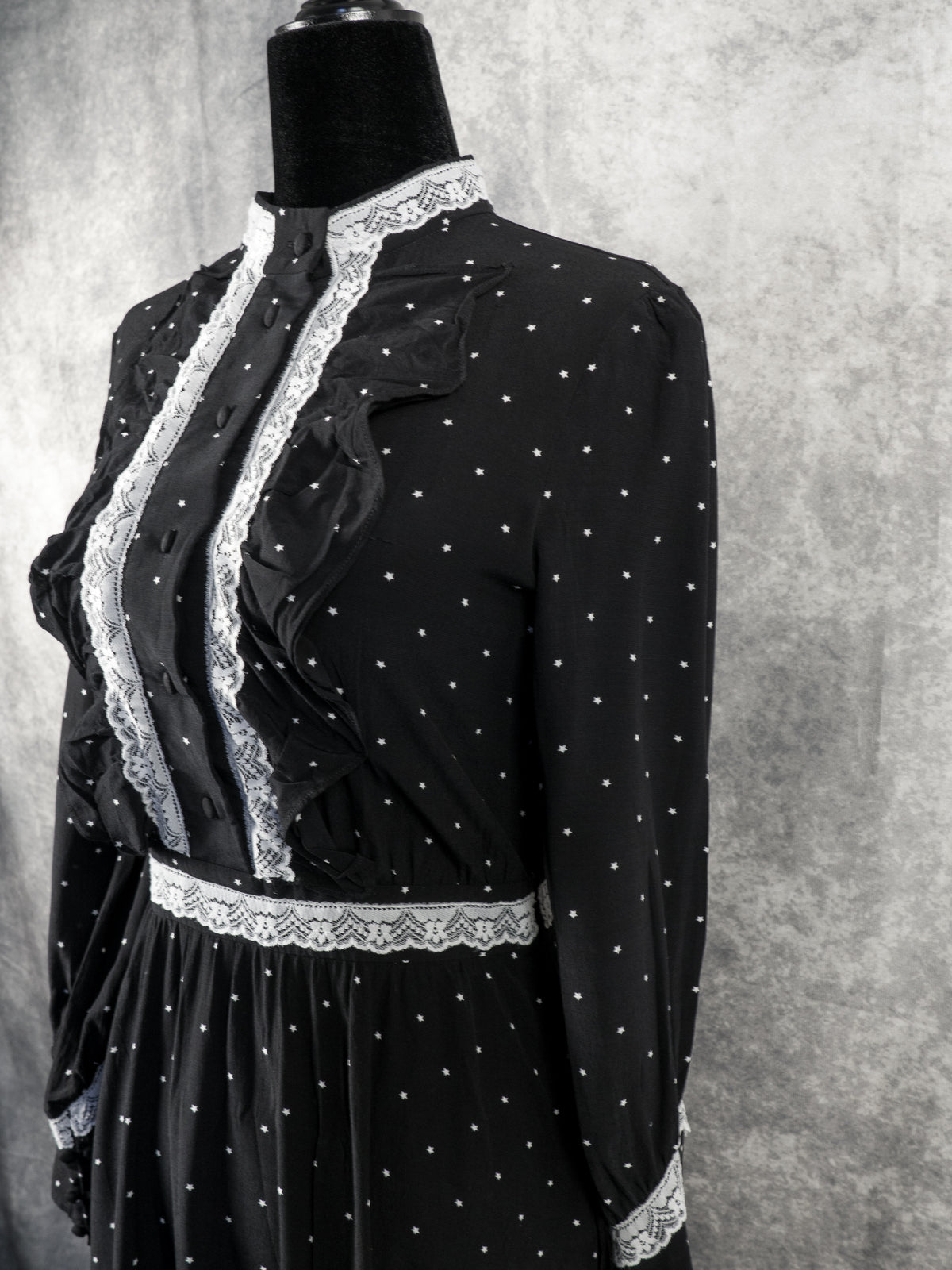 A three-quarter angle view focusing on the upper bodice and torso of the black vintage-style maxi dress, displayed on a mannequin. This shot clearly illustrates how the vertical ruffles and white lace trim flank the buttoned front placket, running from the high collar down to the lace-trimmed waistband. The small white star print repeats across the entire bodice and long sleeves, contrasting against the textured grey backdrop.