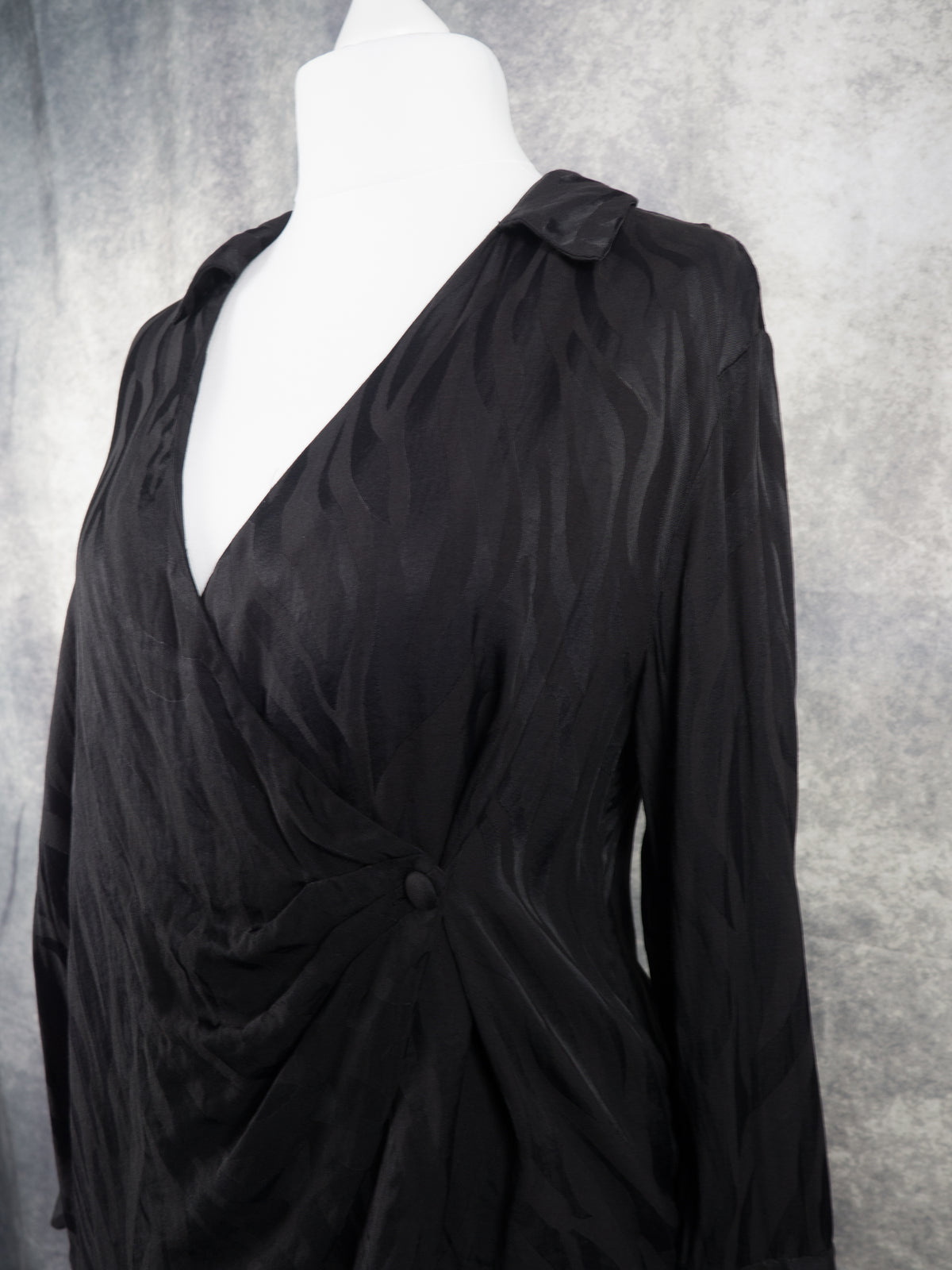 A black long-sleeve wrap-style top with a draped, gathered silhouette, displayed on a white mannequin torso against a textured gray backdrop.