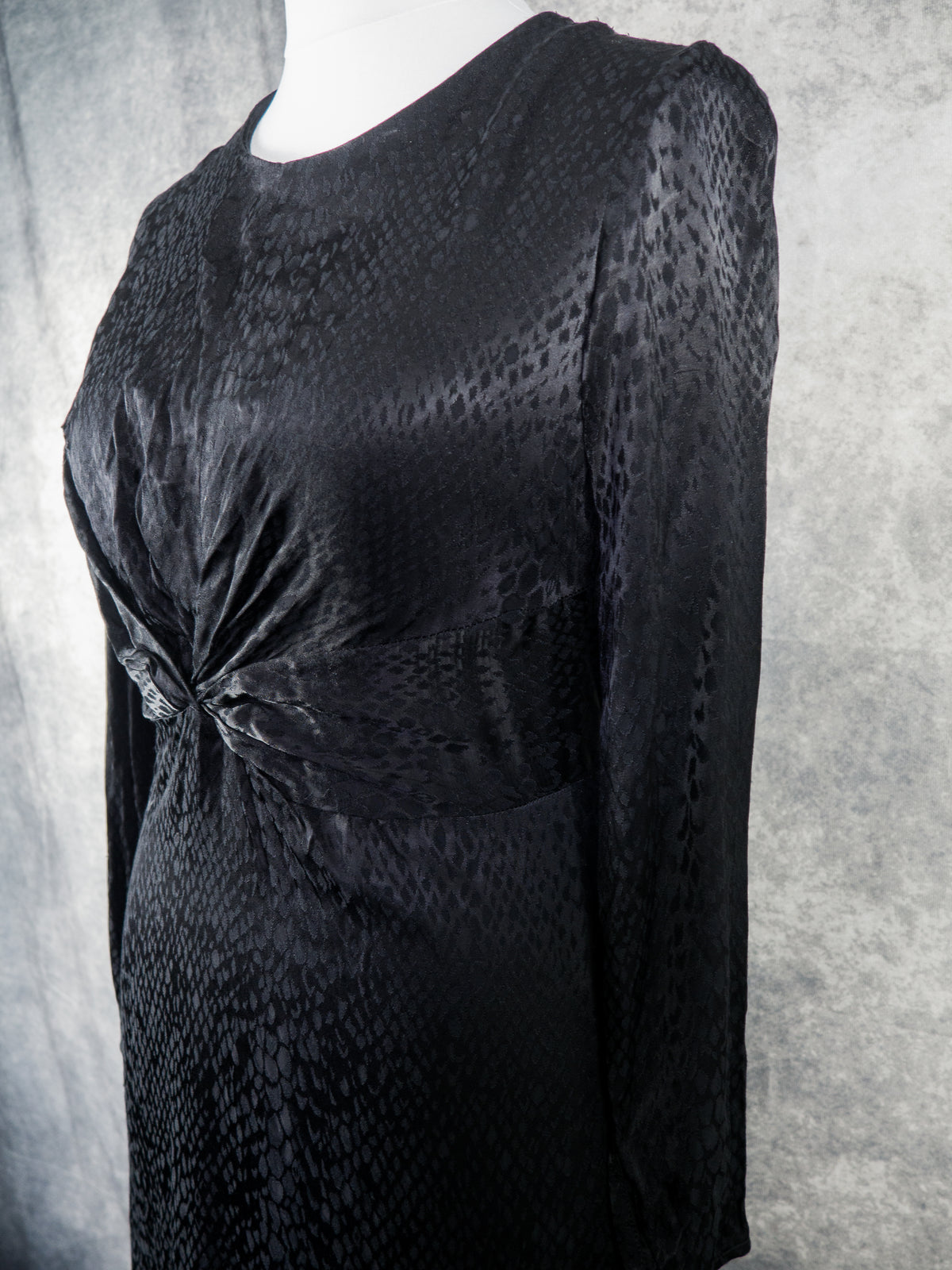 A black long-sleeve dress with a textured, snakeskin-like pattern, displayed on a mannequin against a gray textured backdrop.
