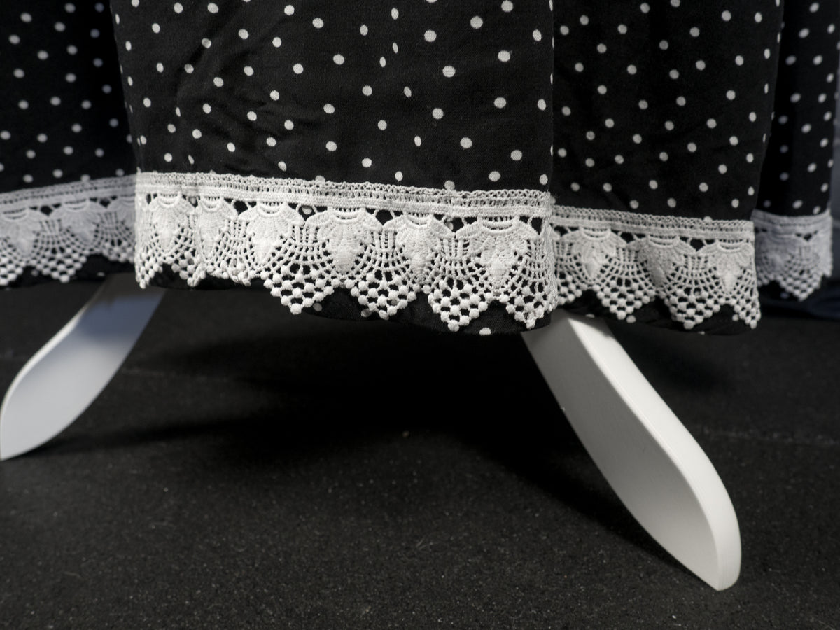 Close-up of the bottom hem of a black dress with a white polka dot pattern. The hem is lined with a broad, intricate white crochet lace trim featuring a scalloped edge. The white wooden legs of a mannequin stand are visible underneath against a dark floor.