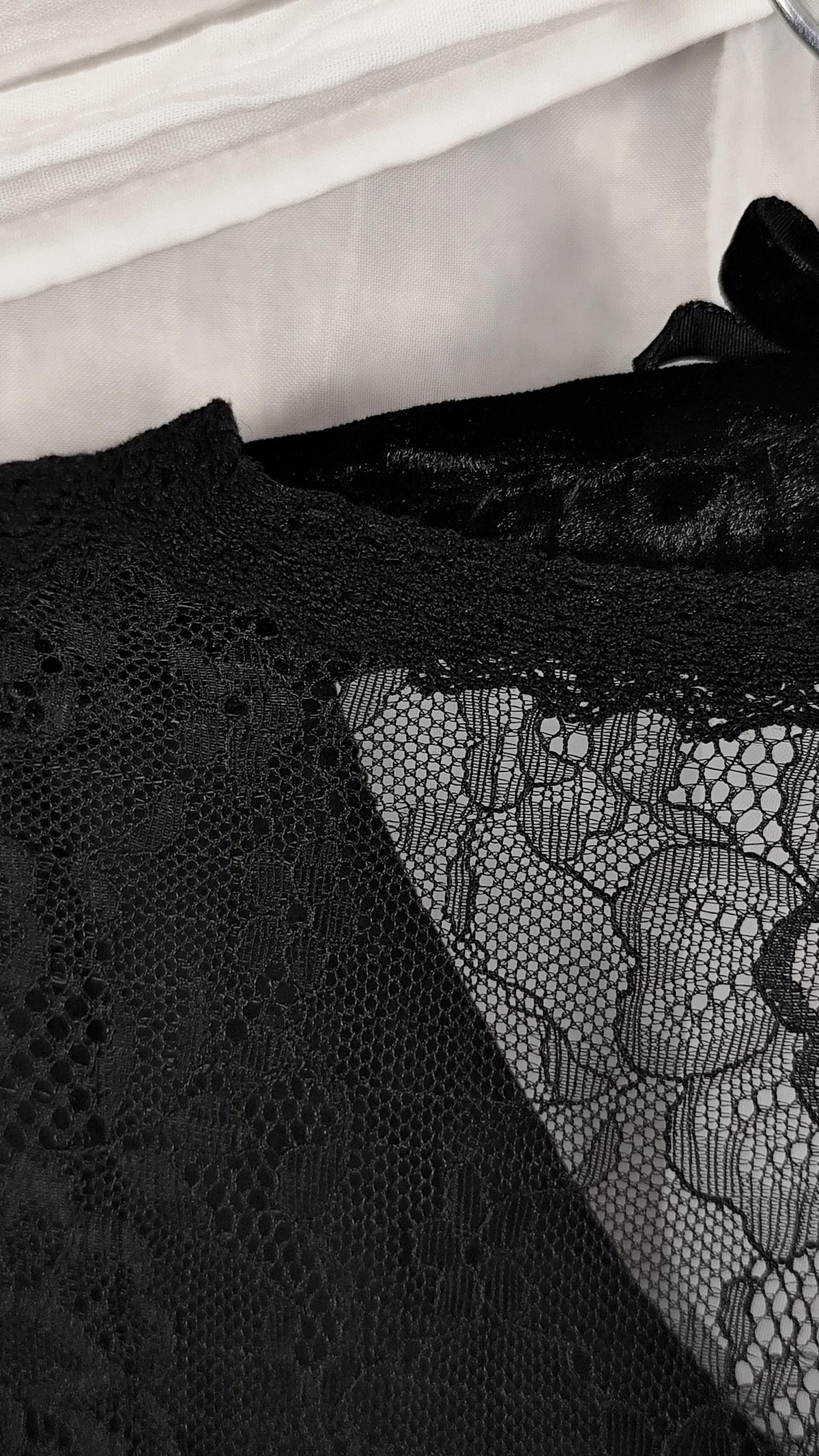Close-up of a black lace garment with sheer floral lace panels and a textured black fabric section, photographed against a light neutral background.