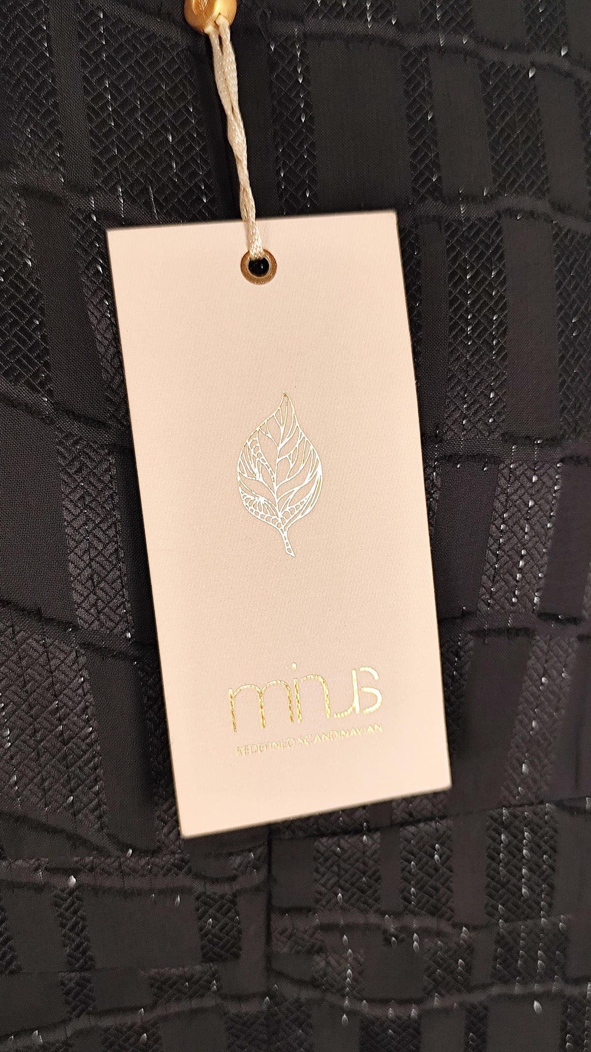 A close-up of a black textured dress with a cream paper hangtag attached by a string. The tag has a metallic leaf logo and the brand name 'Minus' printed in gold at the bottom.