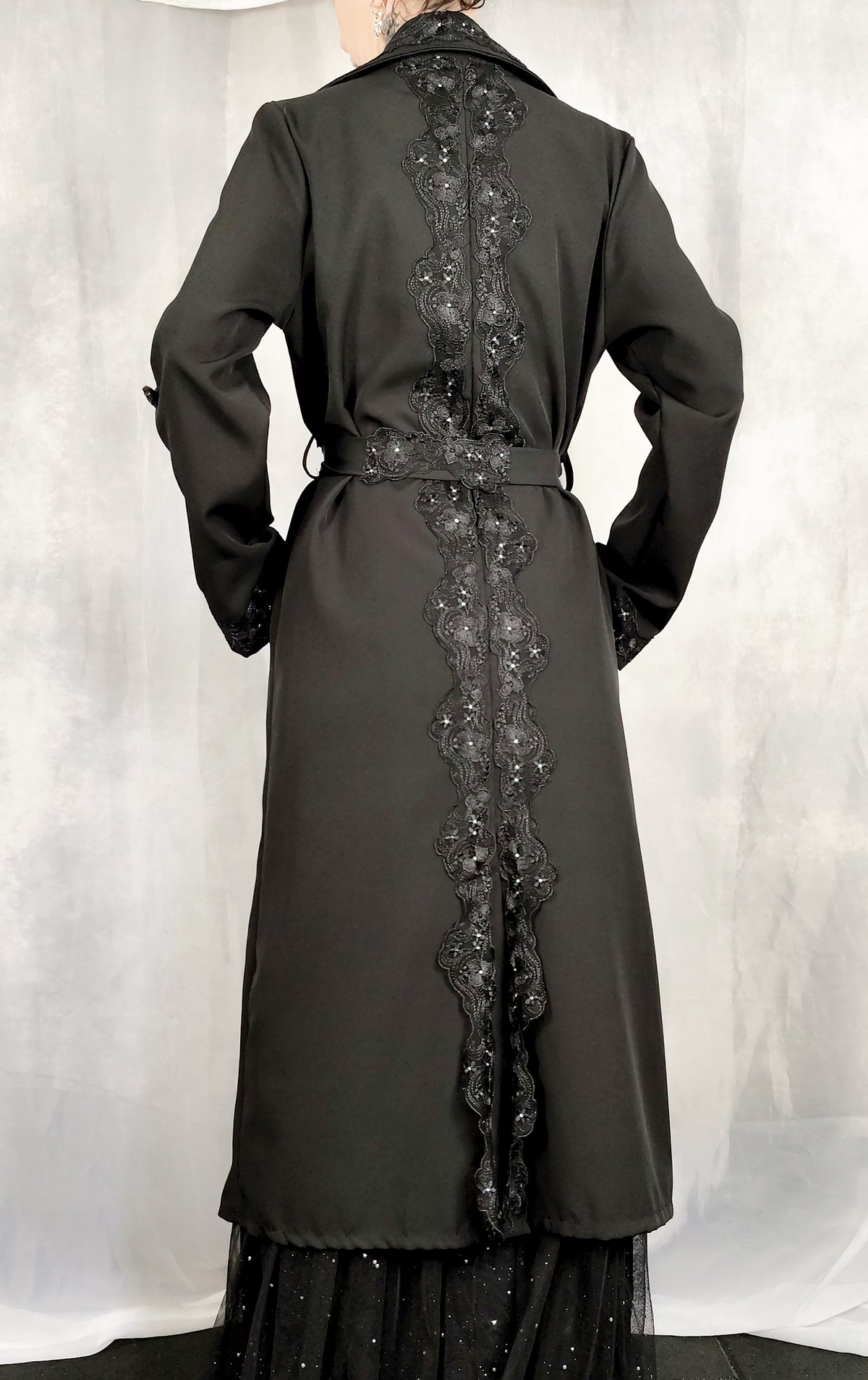 A black long coat with a matching belt and a prominent vertical lace applique detail down the back, shown on a mannequin against a light gray draped backdrop.