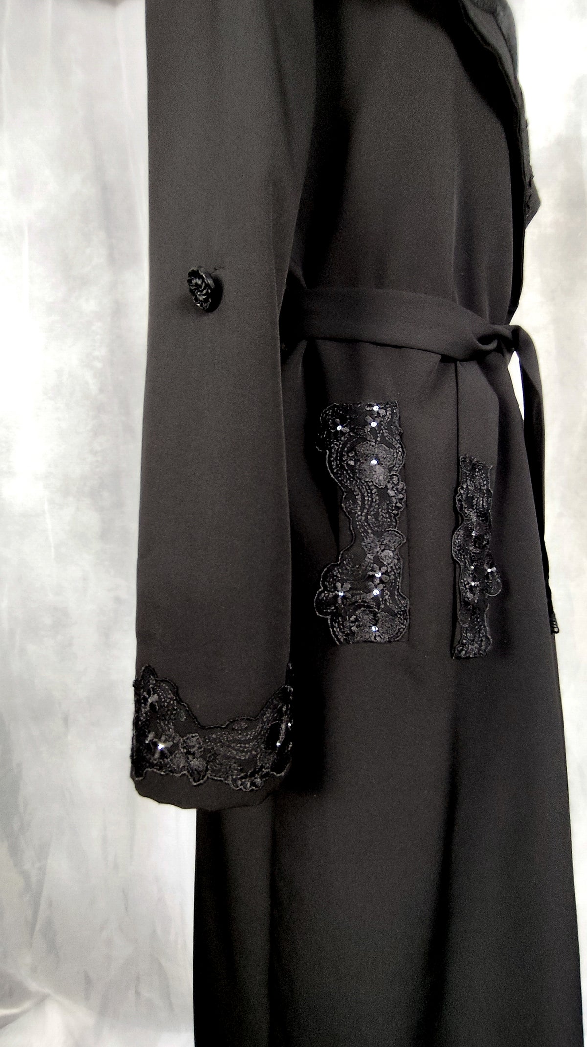 Close-up of long sleeve, pocket and belt of black trench coat, trimmed with lace.