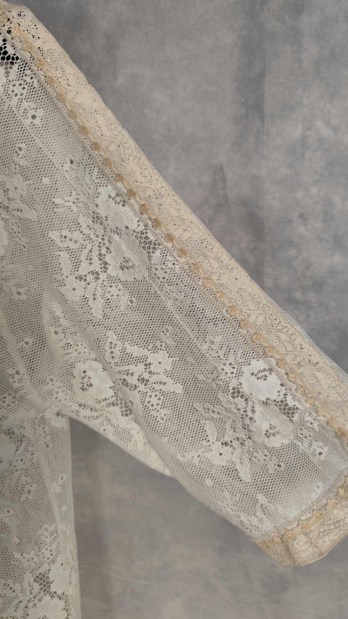Close-up of a pearl-colored lace garment sleeve with floral embroidery and a matching lace cuff, set against a textured gray background. It´s visible a "scar" on the fabric.