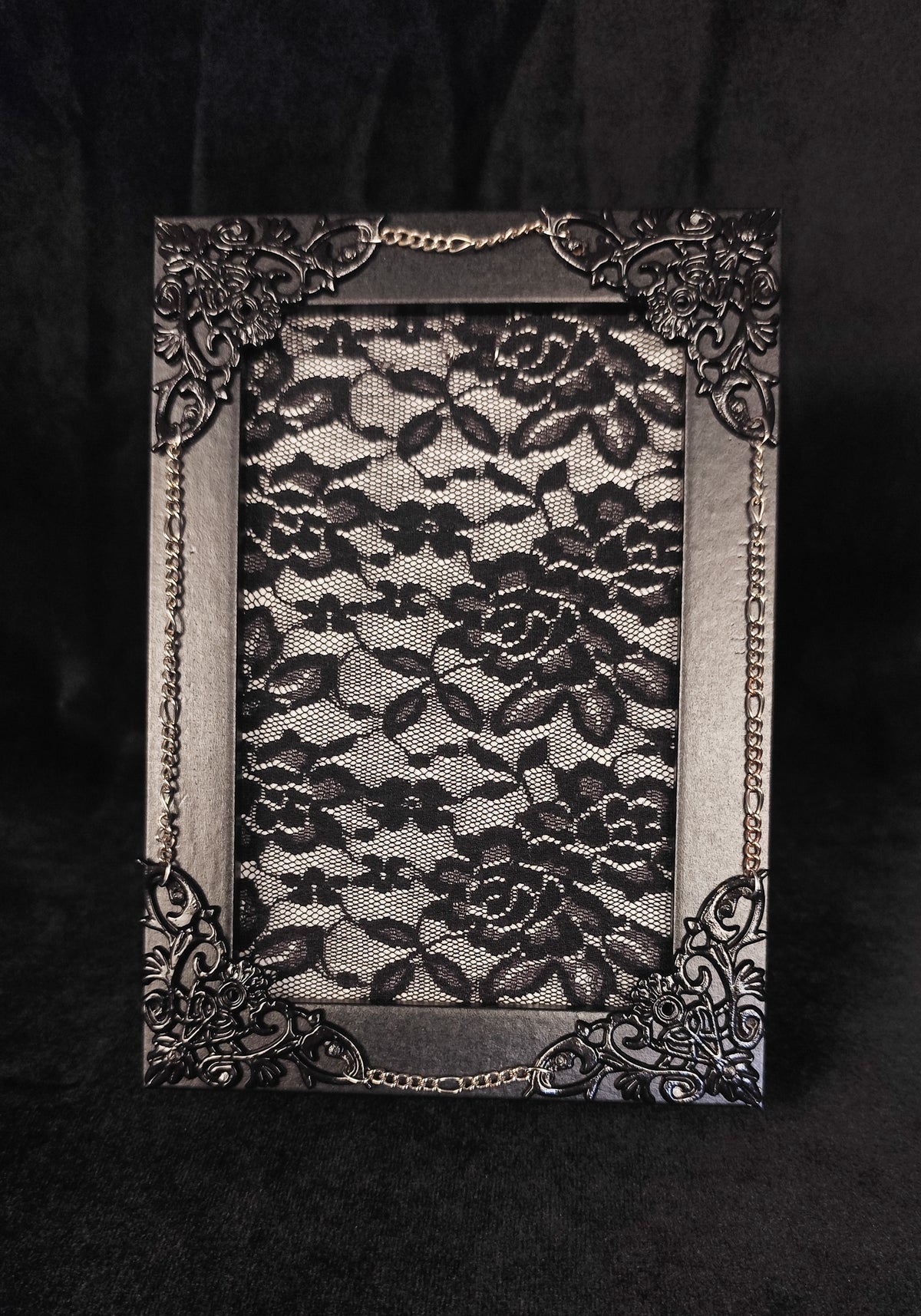 A rectangular black lace picture frame with ornate filigree corners and small chain details, displayed against a dark black fabric background.