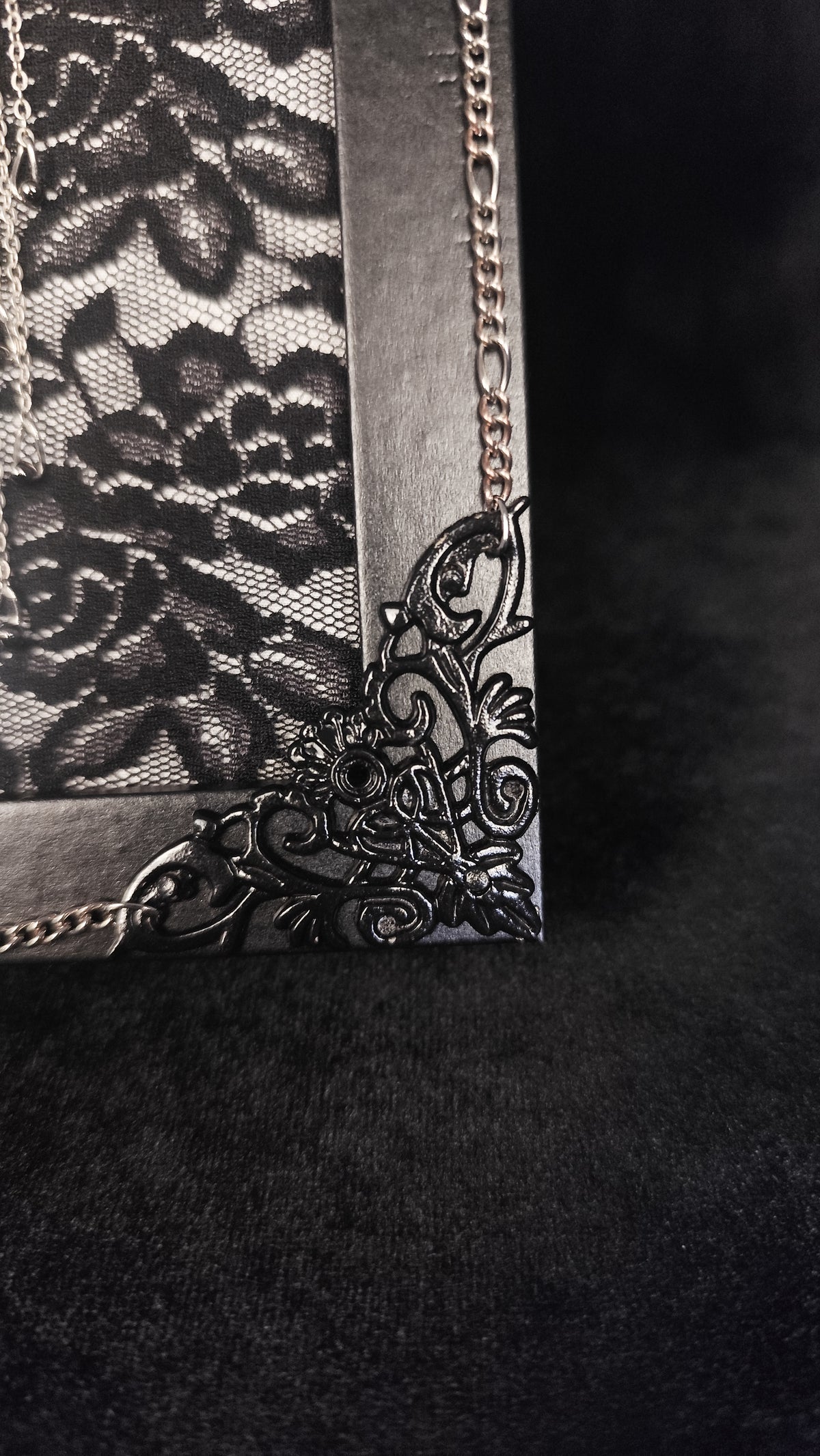 A close-up of a black gothic-style frame corner, featuring a floral lace panel, ornate filigree corner embellishment, and a silver-tone chain strap on a dark textured background.