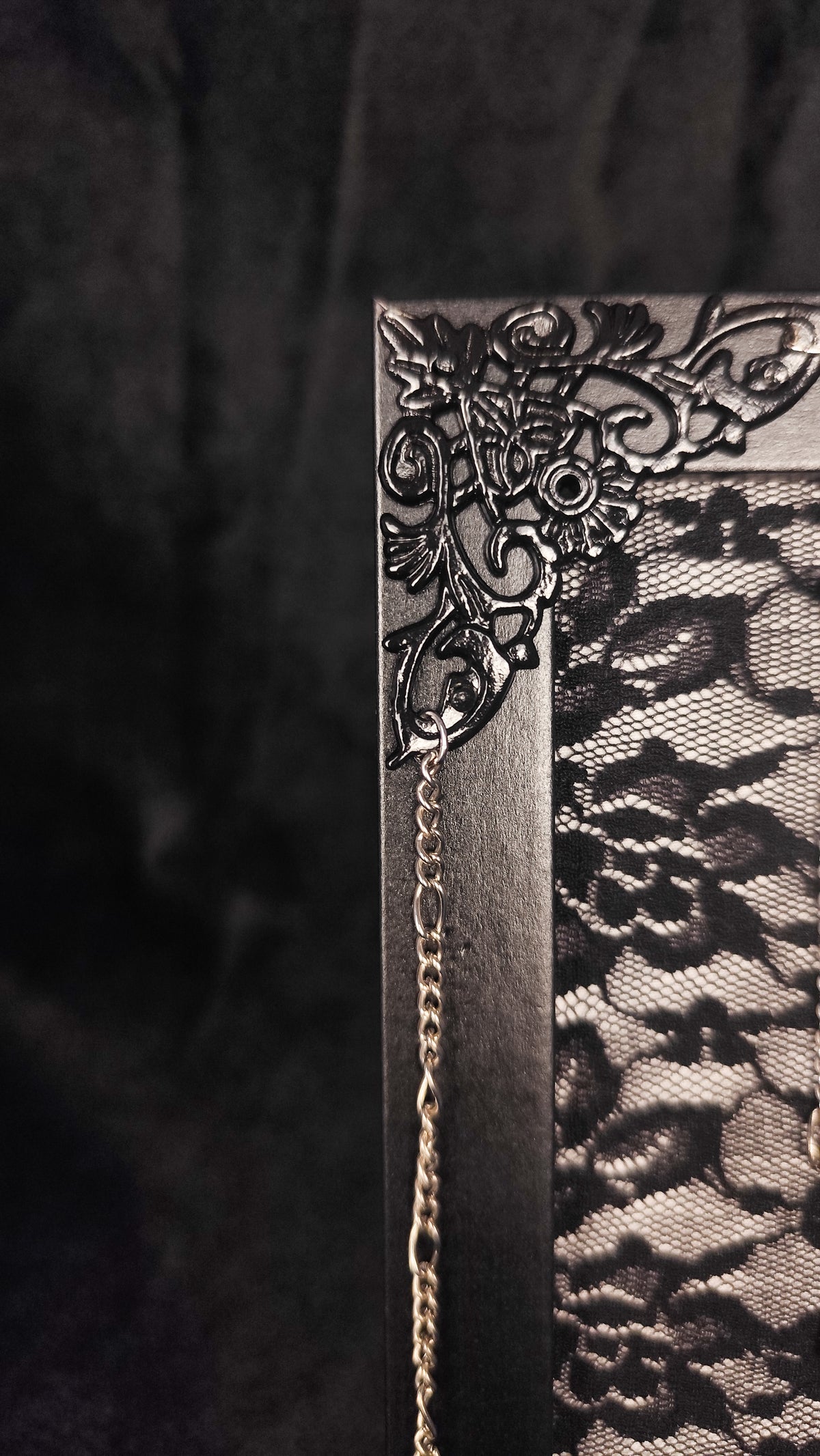 A close-up of a black gothic-style frame with ornate floral corner detailing, a decorative chain, and a black lace cover, set against a dark textured background.