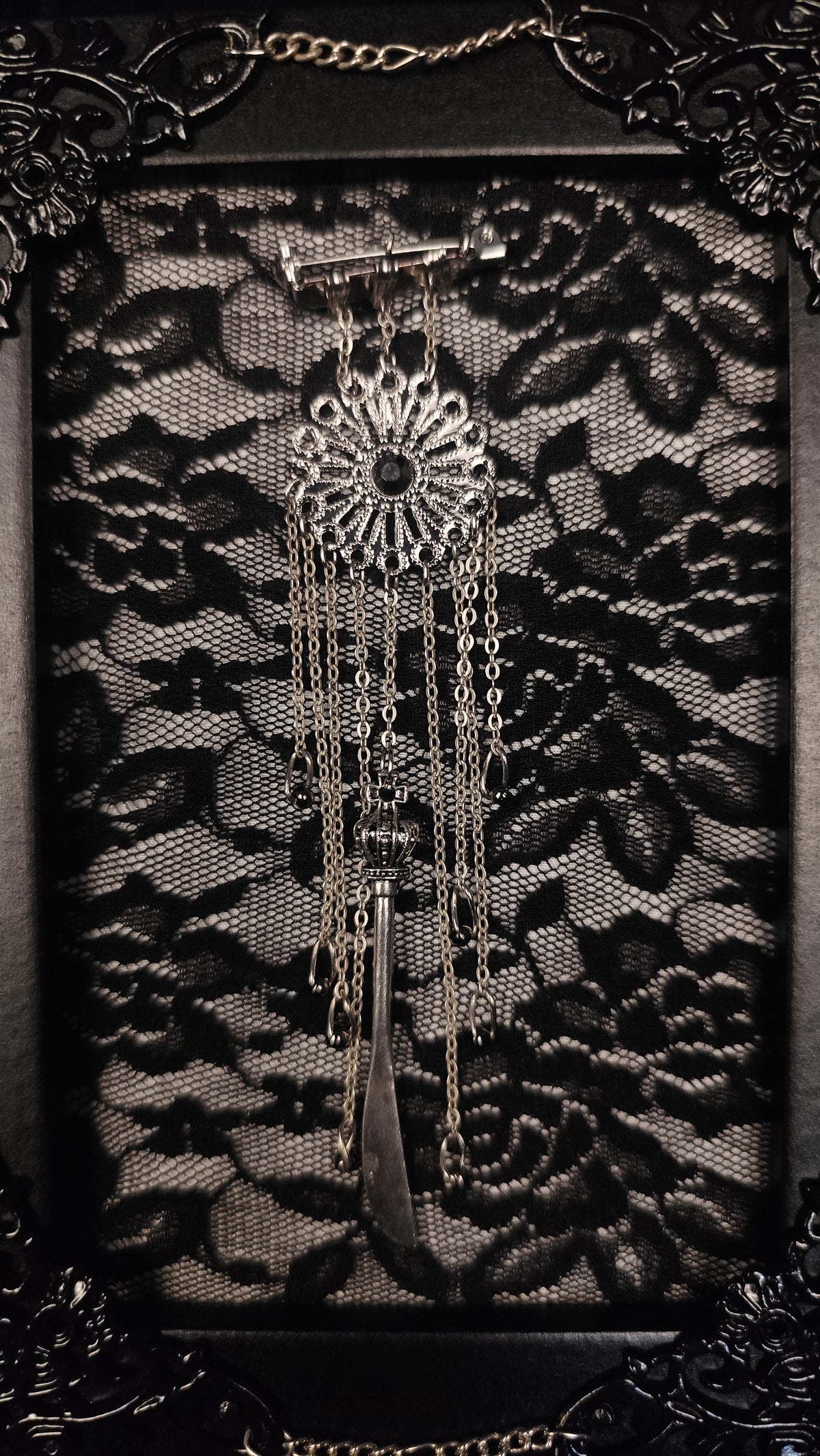 An ornate silver-toned metal table frame with a floral medallion, chains, and a curved blade-like pendant, displayed against a black lace background with a decorative dark frame.