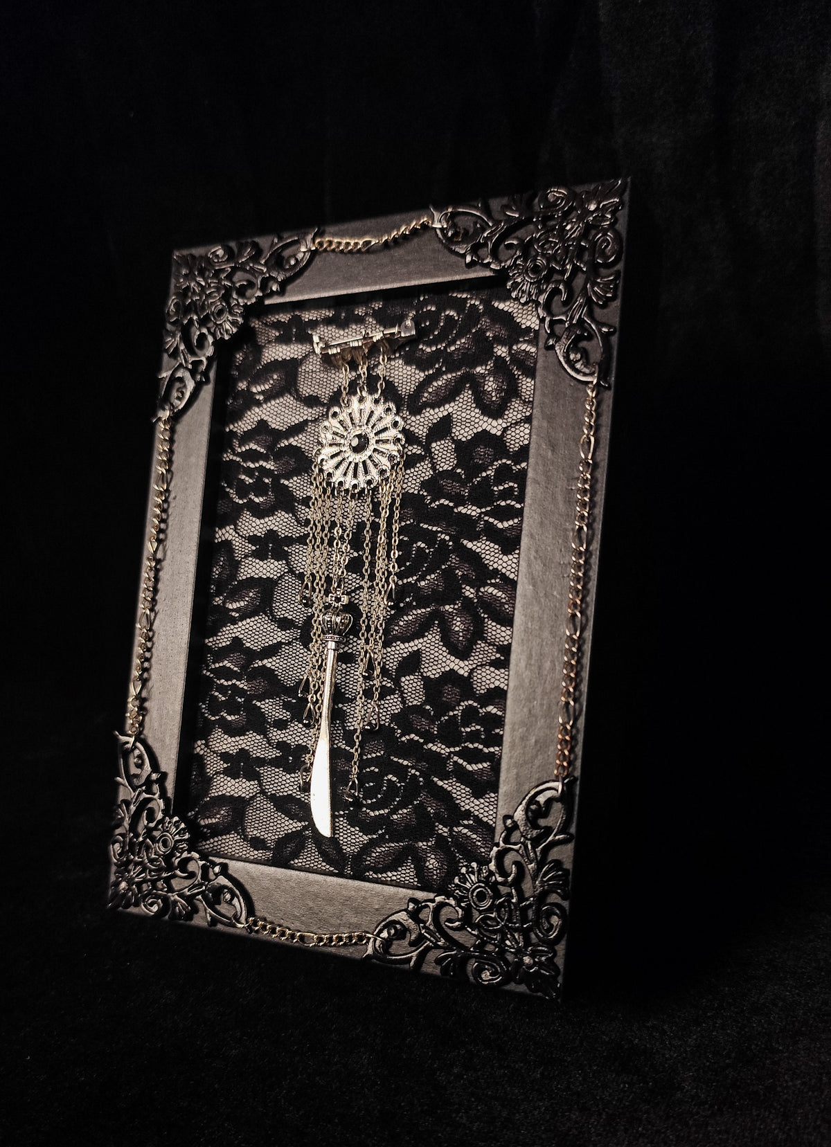 A gothic-style decorative table frame featuring a black lace panel set within an ornate dark metal frame, with a silver dreamcatcher and white tassel suspended in the center against a black background.