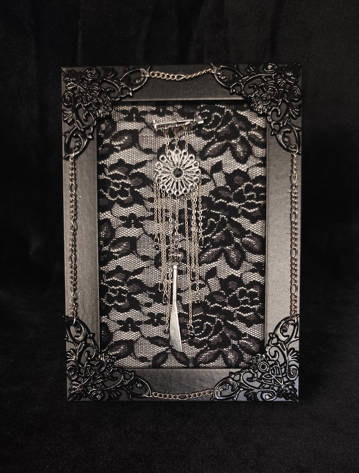 A gothic-style framed table art piece featuring a black lace background, an ornate black filigree frame with silver-toned chain accents, and a silver dreamcatcher hanging in the center, set against a dark black fabric backdrop.