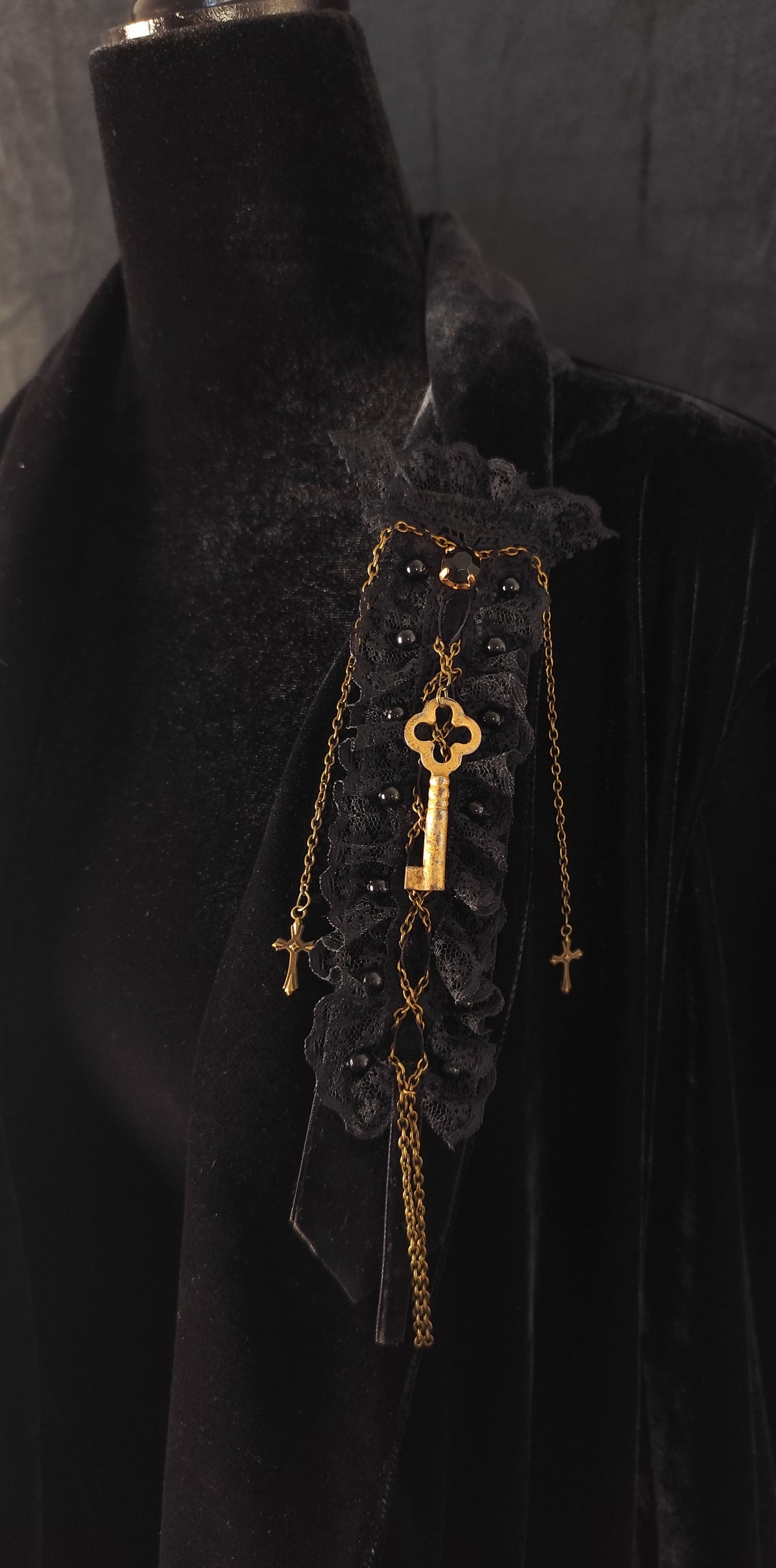 A gothic-style black lace brooch with a gold key, keychain, and cross details, displayed on a black velvet mannequin torso.