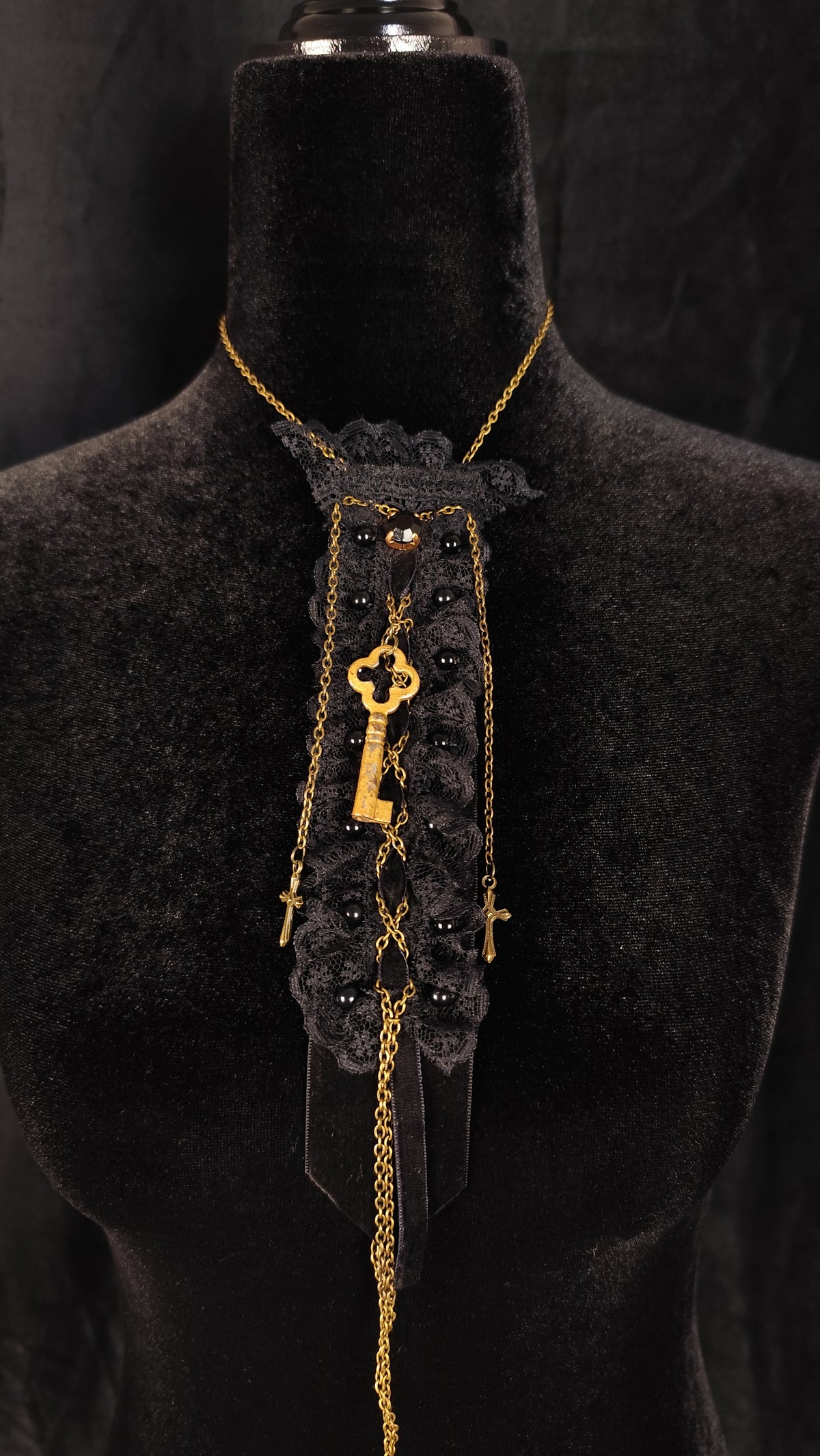 A gothic-style necklace displayed on a black velvet mannequin bust. The necklace features a black lace collar with a central gold key charm, black beads, and gold chains.