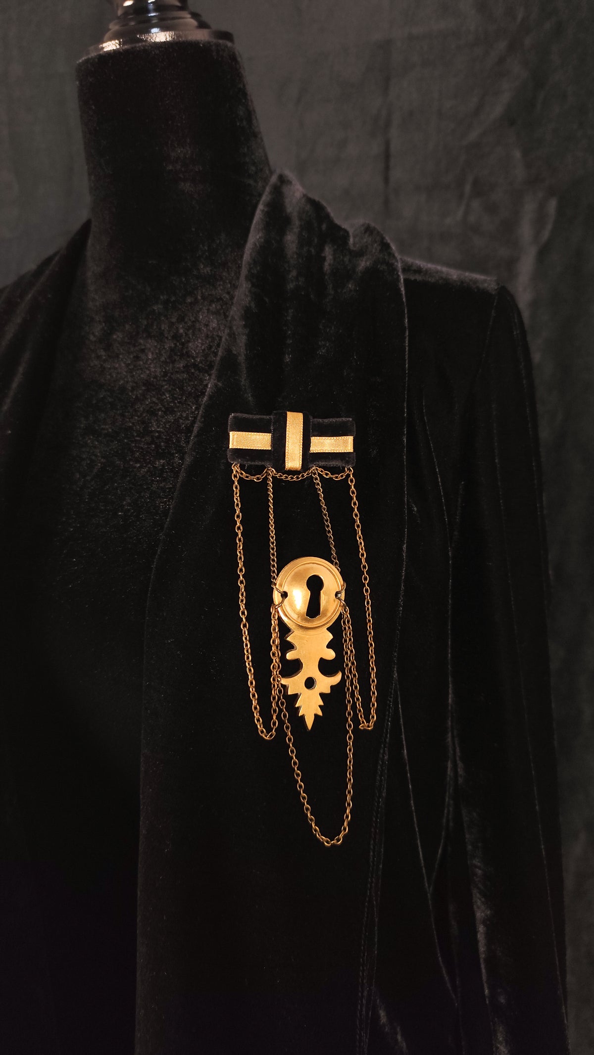 A gold-toned gothic keyhole brooch with black enamel details, displayed on a black velvet mannequin torso against a dark textured background.