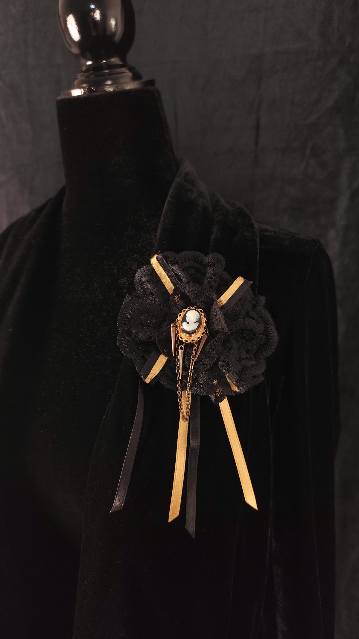 A black lace brooch with gold ribbon accents and a central oval cameo, displayed on a black velvet mannequin torso against a dark background.