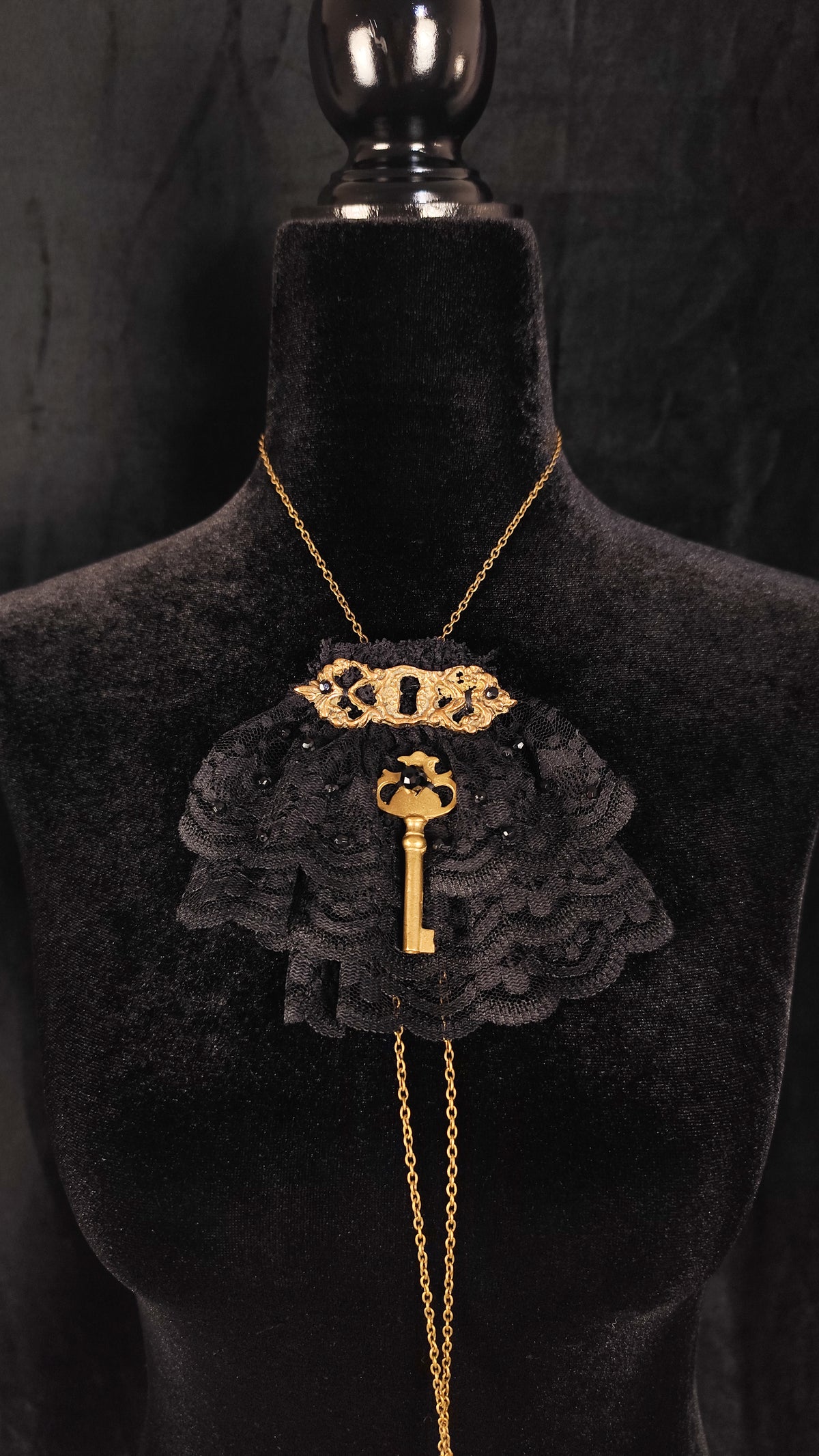 A gothic-style necklace displayed on a black velvet mannequin bust. The necklace features a gold chain, a large black ruffled lace collar, a gold key pendant, and a gold crest at the collarband.