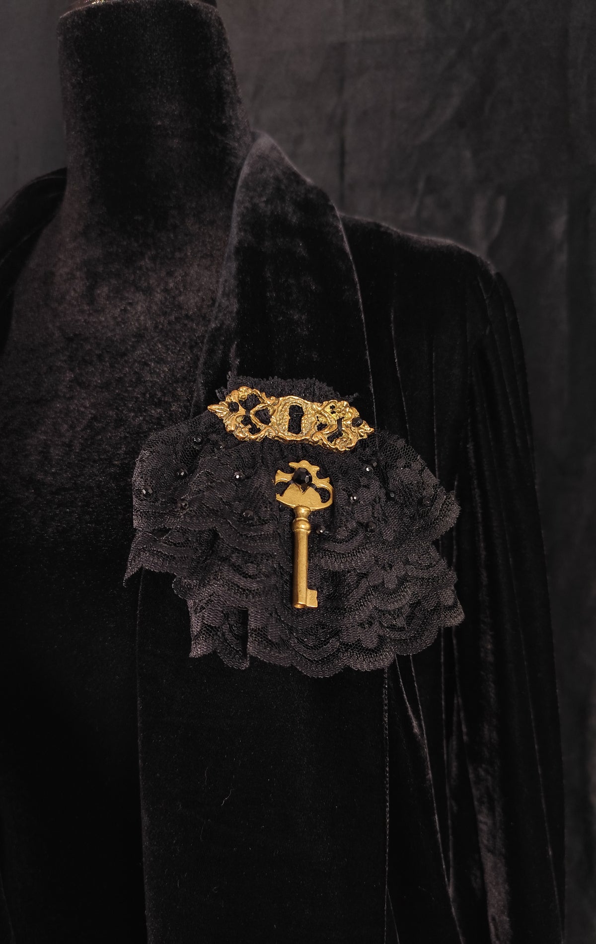 A black velvet and lace brooch featuring a gold key and keyhole design, displayed on a black velvet mannequin torso against a dark textured background.