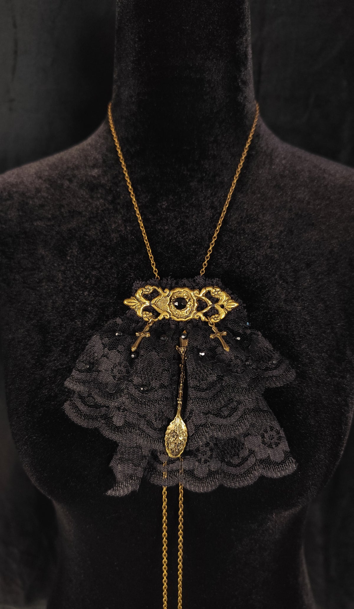 A gothic-style necklace displayed on a black velvet mannequin bust. The necklace features a gold-tone chain, a central gold-tone vintage keyhole, and a black lace ruffled collar with a dangling gold-tone spoon charm.