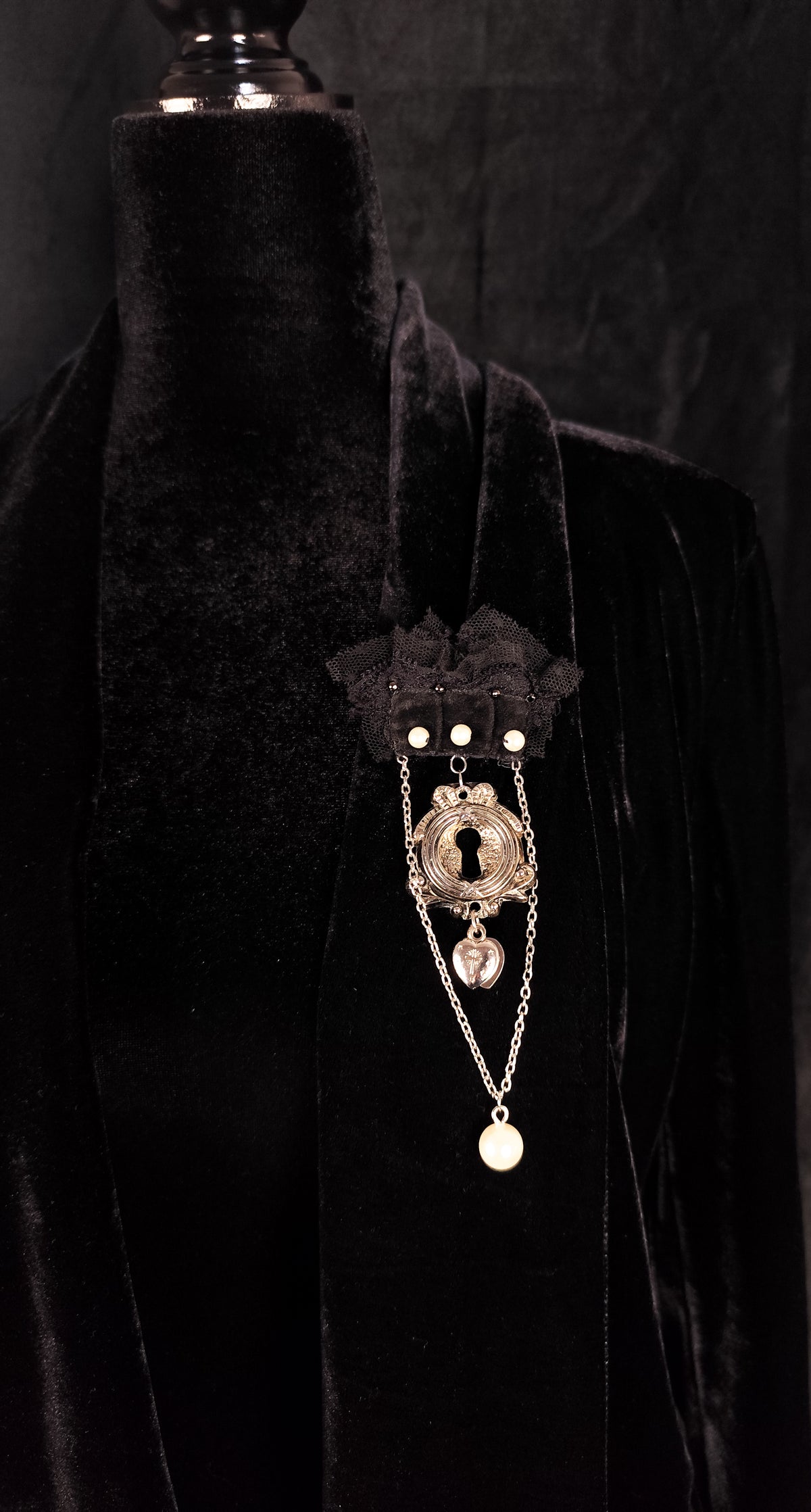 A gothic-style brooch featuring a silver-toned keyhole pendant, and pearl accents, displayed on a black velvet mannequin torso.