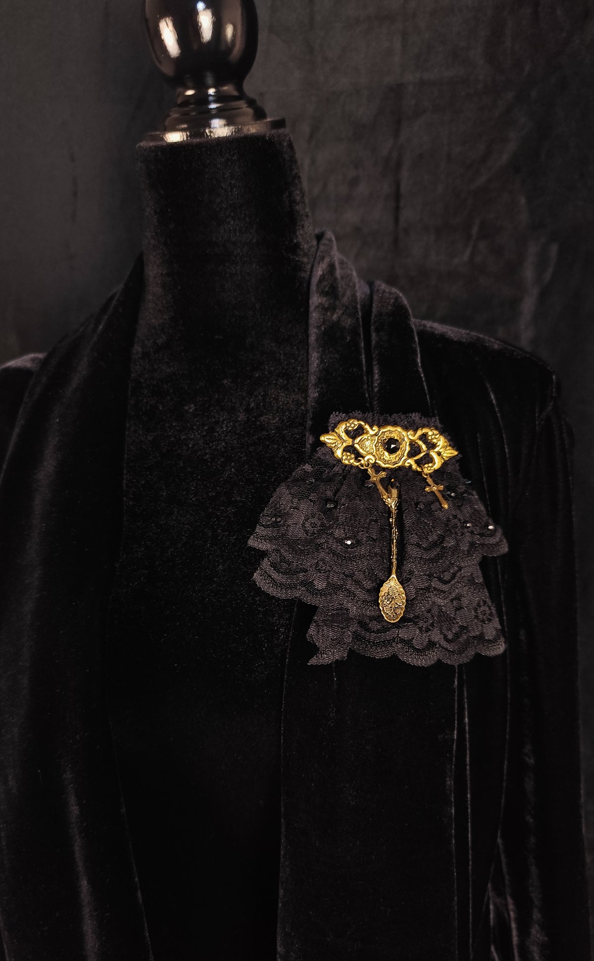 A black lace ruffled brooch with gold filigree and a dangling gold spoon charm, displayed on a black velvet mannequin torso against a dark textured background.