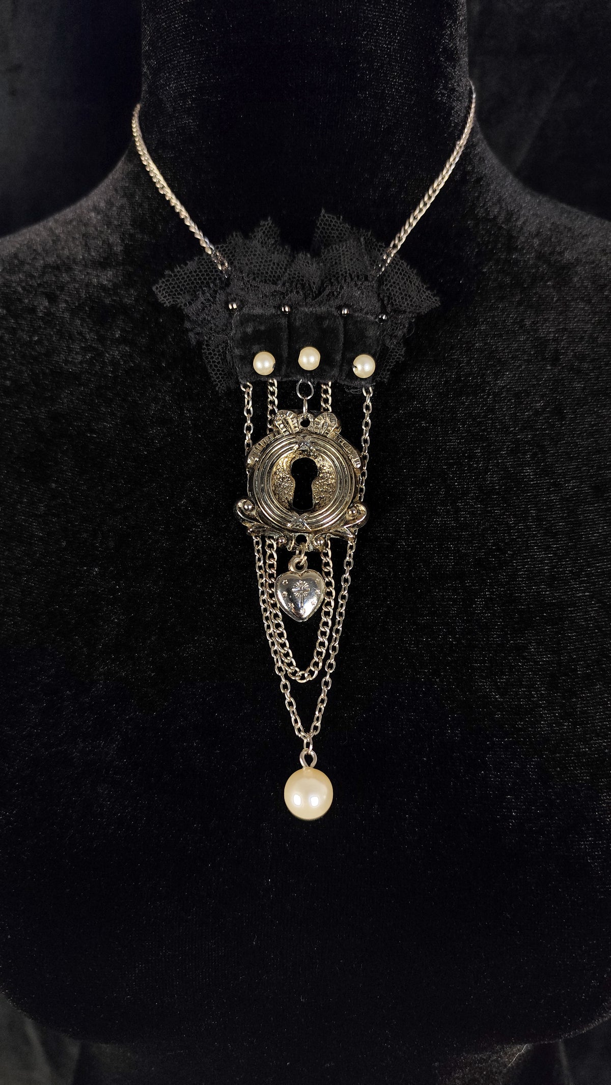A gothic-style gothic necklace displayed on a black velvet mannequin bust. The necklace features a black lace collar, a central antique silver keyhole pendant, small white faux pearls, a clear heart-shaped crystal, and a dangling white pearl bead.