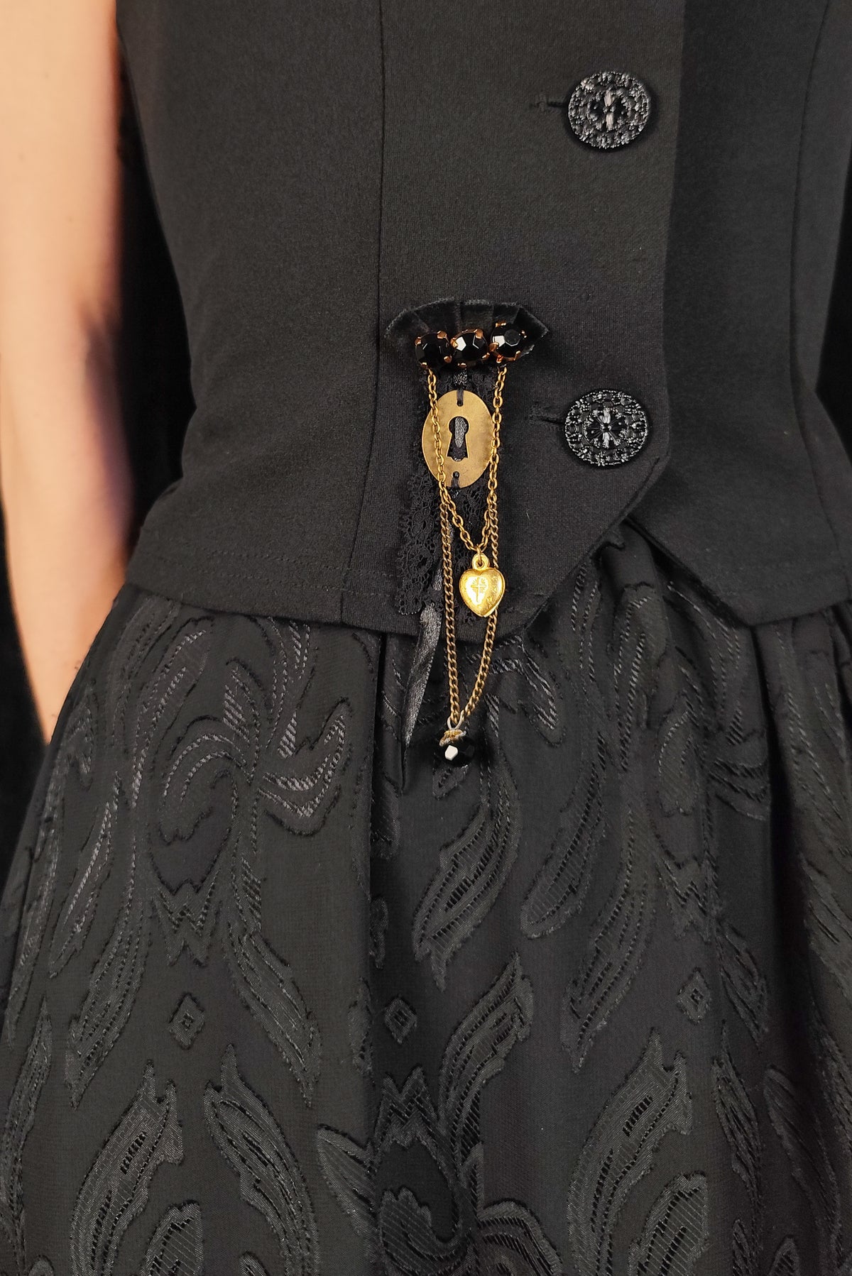 Close-up of a black dress with a structured waist, decorative buttons, and a gold keyhole and heart pendant accessory at the center front.