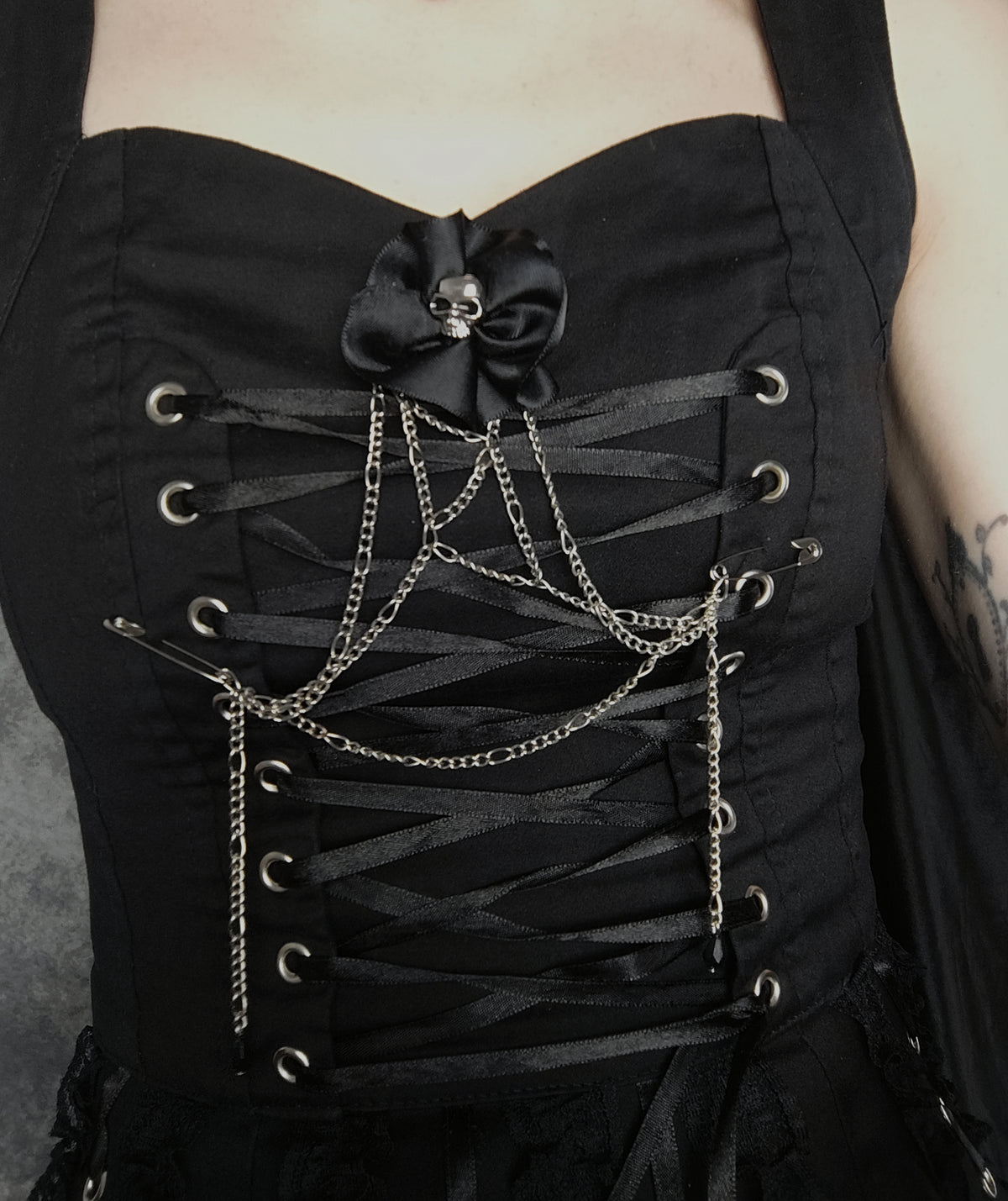 Close-up of a black corset top with ribbon lacing, silver-tone metal eyelets, draped chain details, and a black satin bow with a skull ornament at the center.