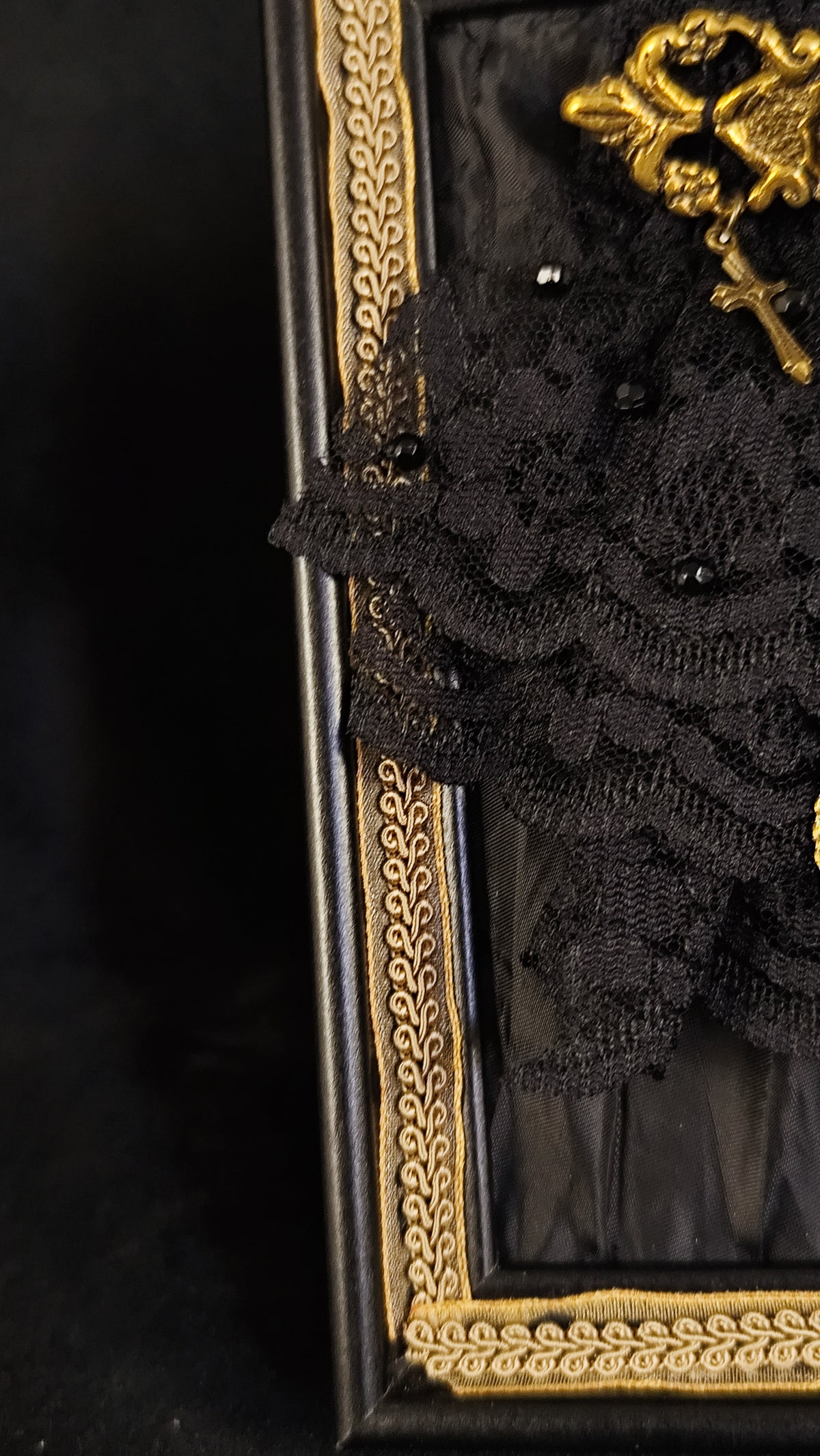 A close-up of a black lace floral arrangement with small black beads and a gold decorative accent, set against a dark background with a black and gold ornate frame visible on the left.