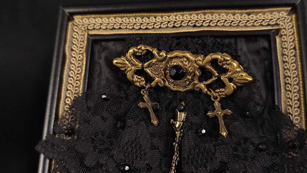 An ornate gold-toned brooch with floral scrollwork and two dangling crosses, displayed on a black lace surface inside a decorative black and gold frame.