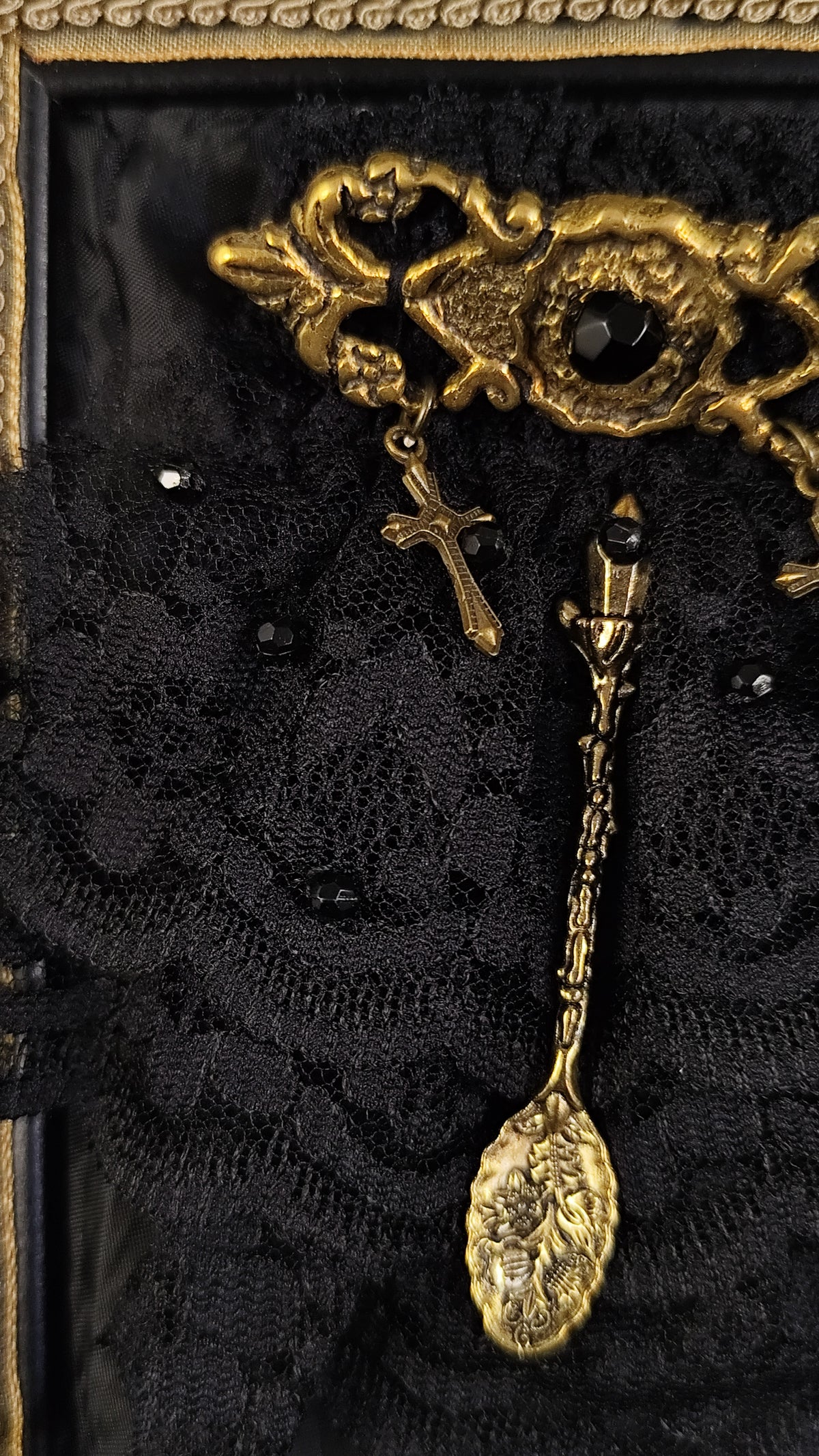 An ornate antique-style gold-toned brooch with a cross charm and a spoon charm, displayed on black lace fabric inside a decorative frame.