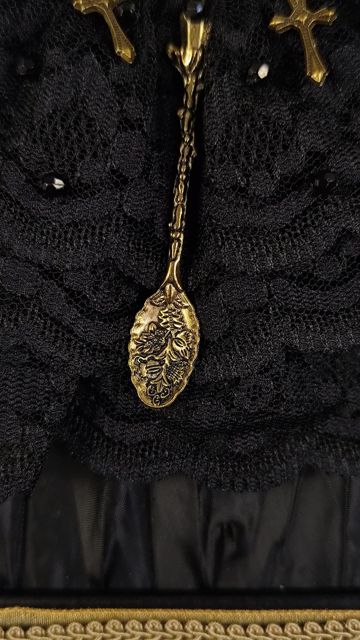 Close-up of a black lace garment with ornate gold metal embellishments, including a decorative hanging piece and cross-shaped details.