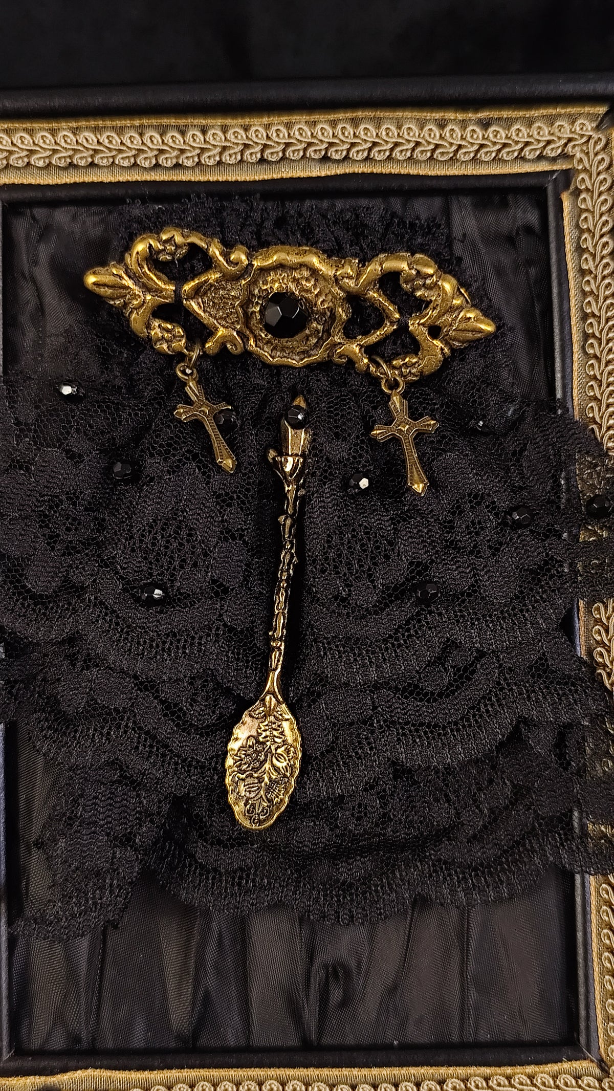 An ornate gold-toned gothic brooch featuring a central oval motif with a black stone, flanked by two dangling crosses, displayed on a black lace background inside a decorative gold frame.