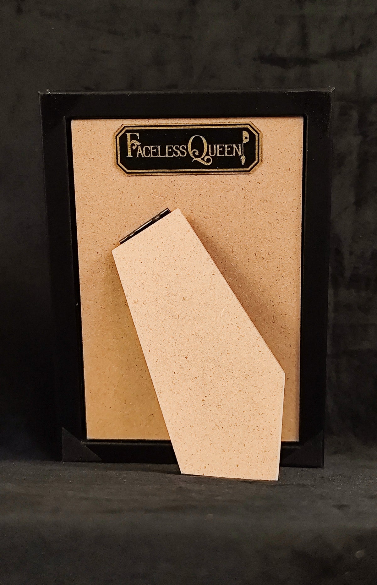 A black-framed picture frame with a beige insert board and a matching beige leaning card, featuring the 'Faceless Queen' logo at the top, set against a dark textured background.