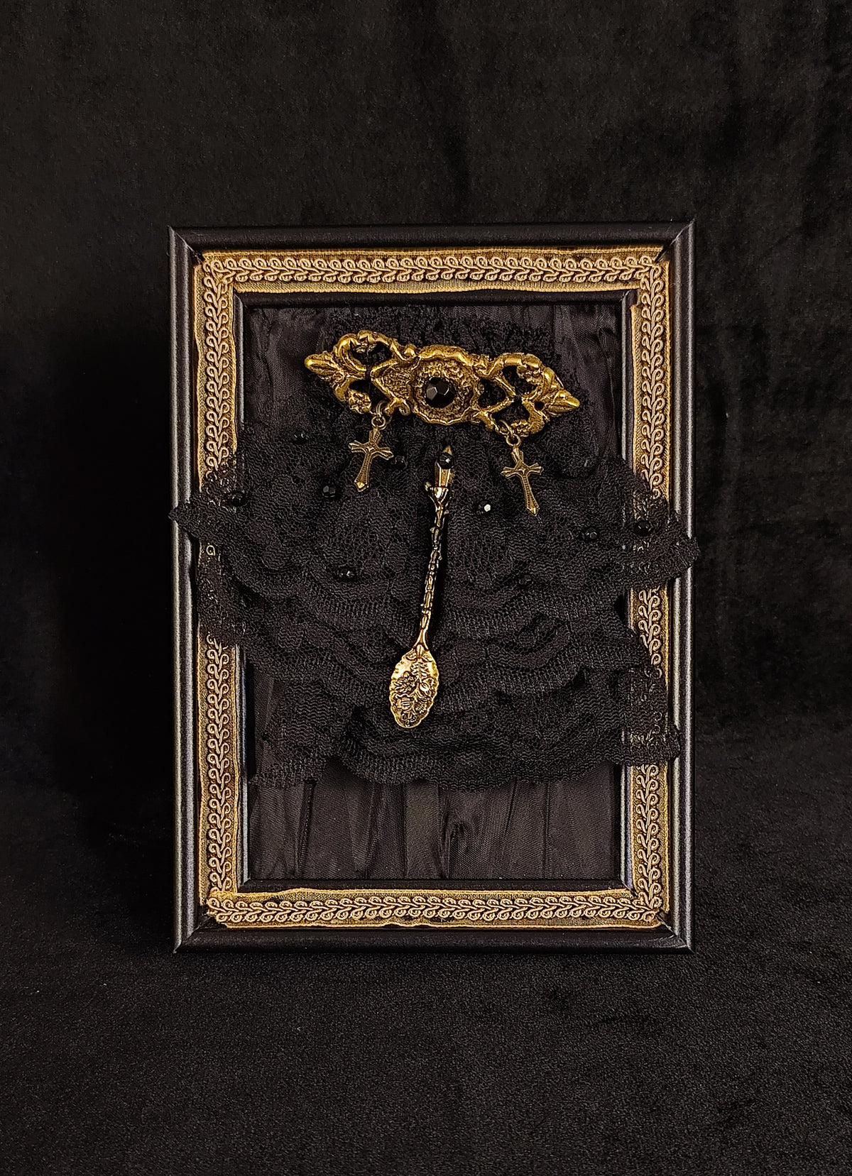 A framed gothic table art piece featuring a black lace ruffled backdrop, a gold ornate brooch with a central keyhole, and a dangling gold spoon charm, all set in a black frame with an ornate gold border.