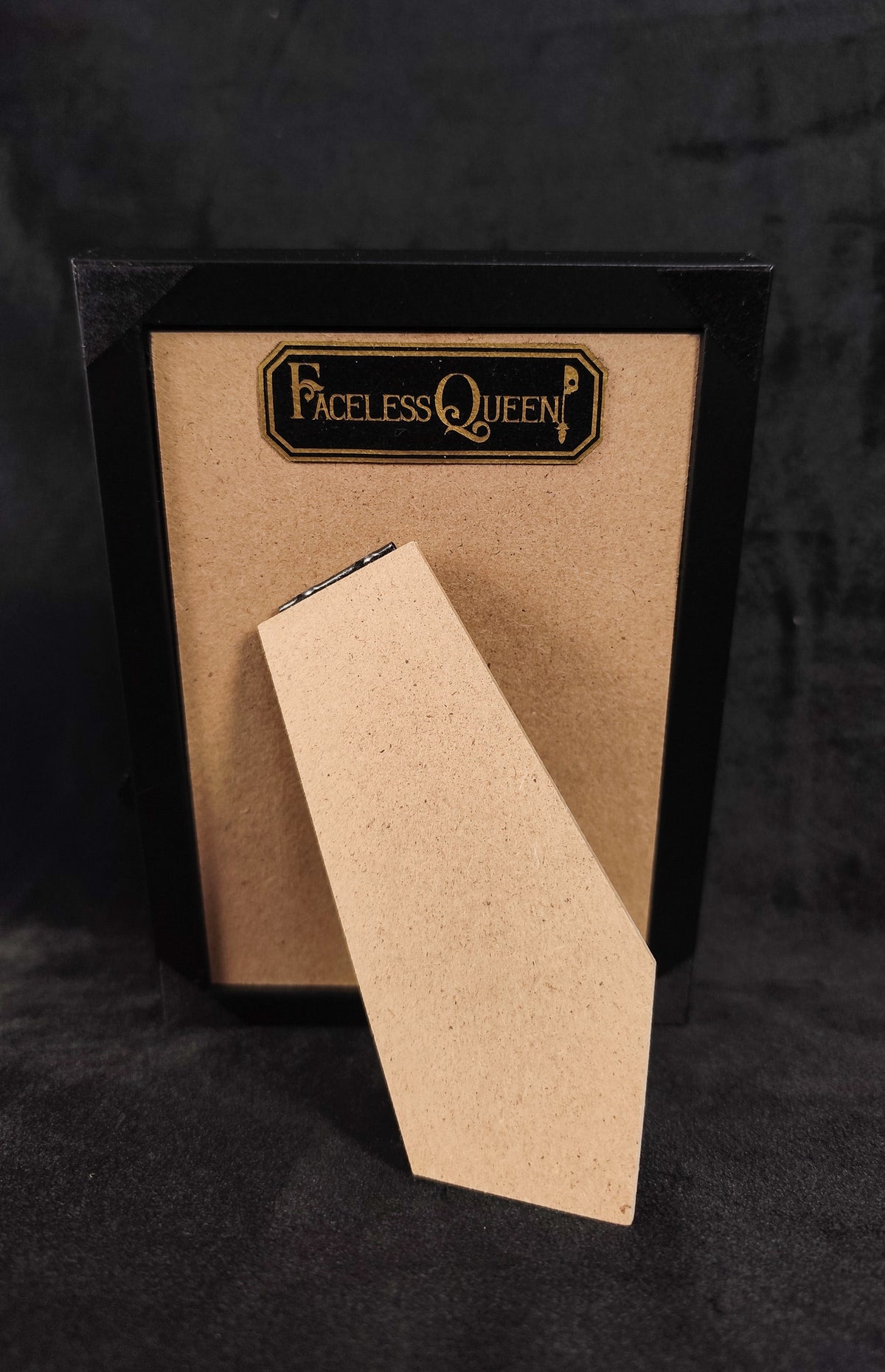 A black-framed picture frame with a tan textured insert and a matching leaning card, branded with a gold 'Faceless Queen' logo, set against a dark textured background.