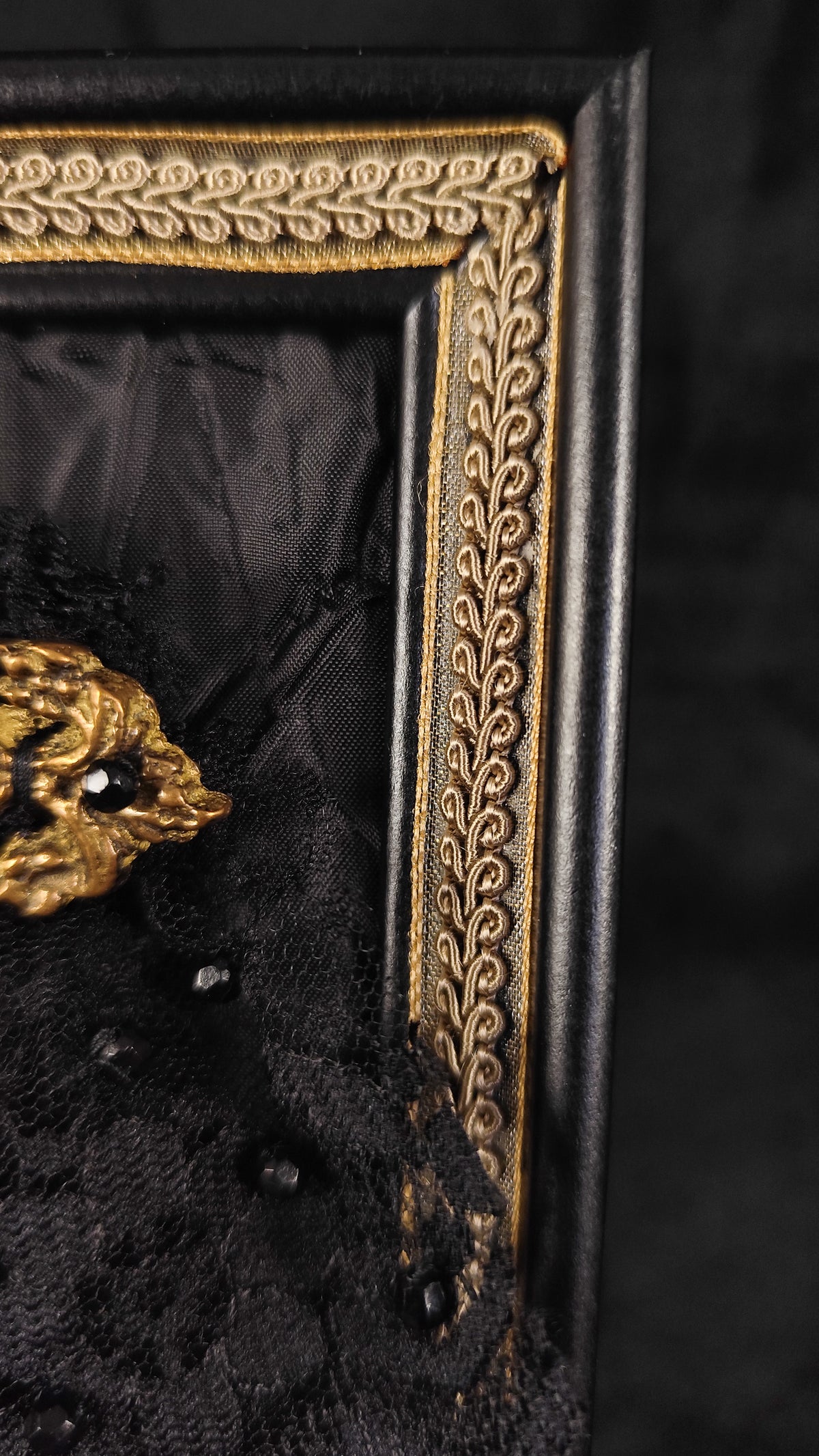 Close-up of a black and gold decorative frame with ornate gold embroidery, black lace fabric, and a small gold embellishment on a dark background.