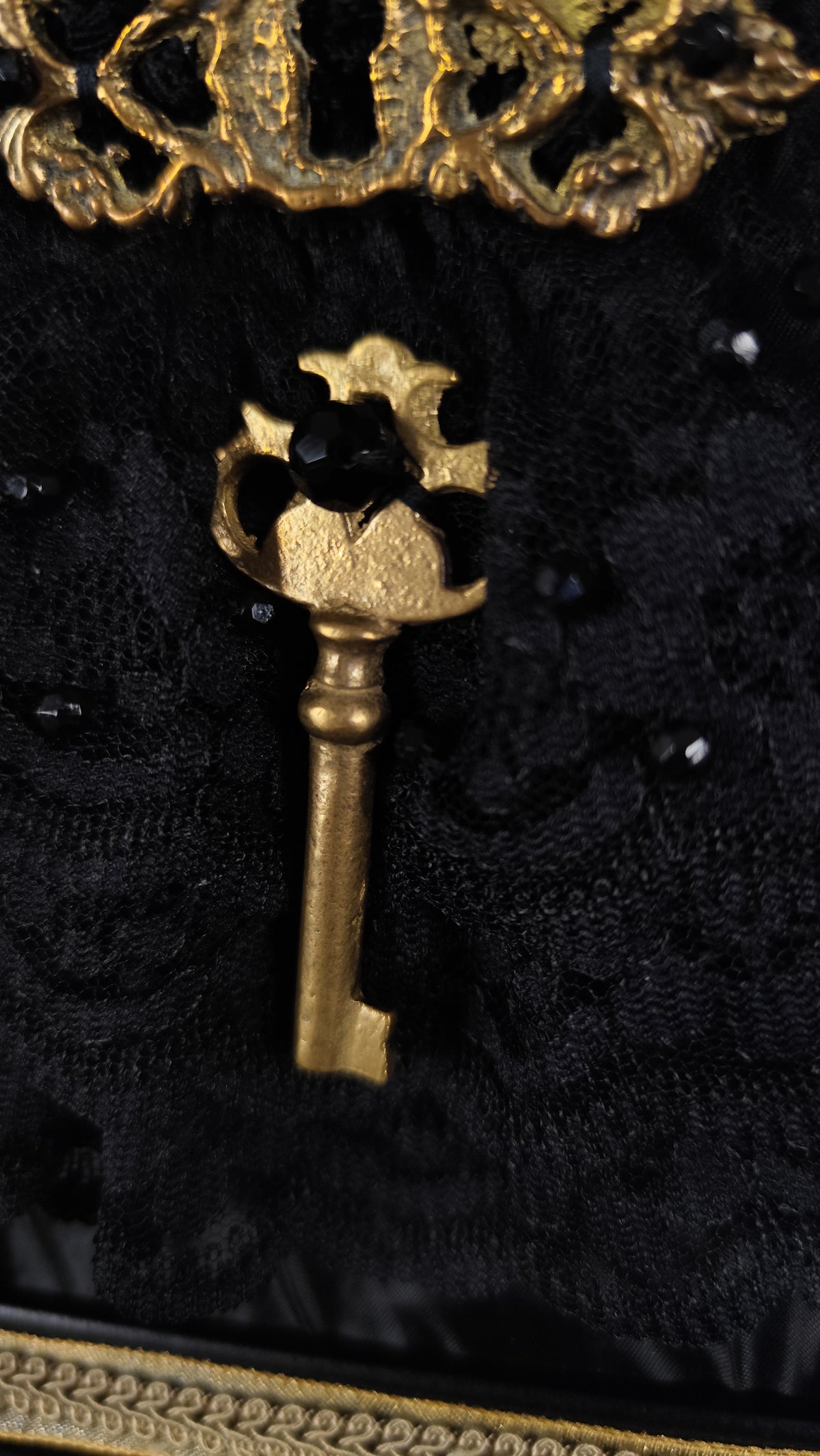 A vintage-style gold key with a decorative crown-like top, displayed against a black lace fabric background with small black beads.