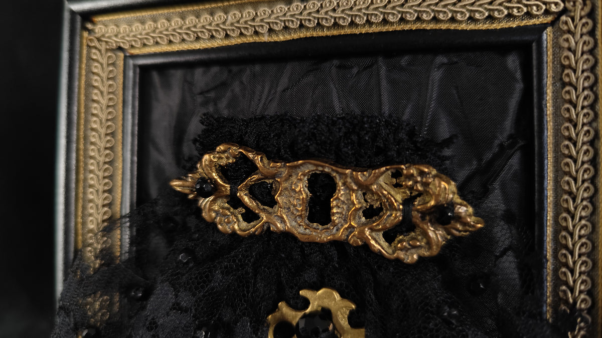 A gothic-style decorative accessory featuring an ornate gold keyhole-shaped centerpiece with a keyhole cutout, layered over black lace and tulle, displayed inside a black and gold frame.