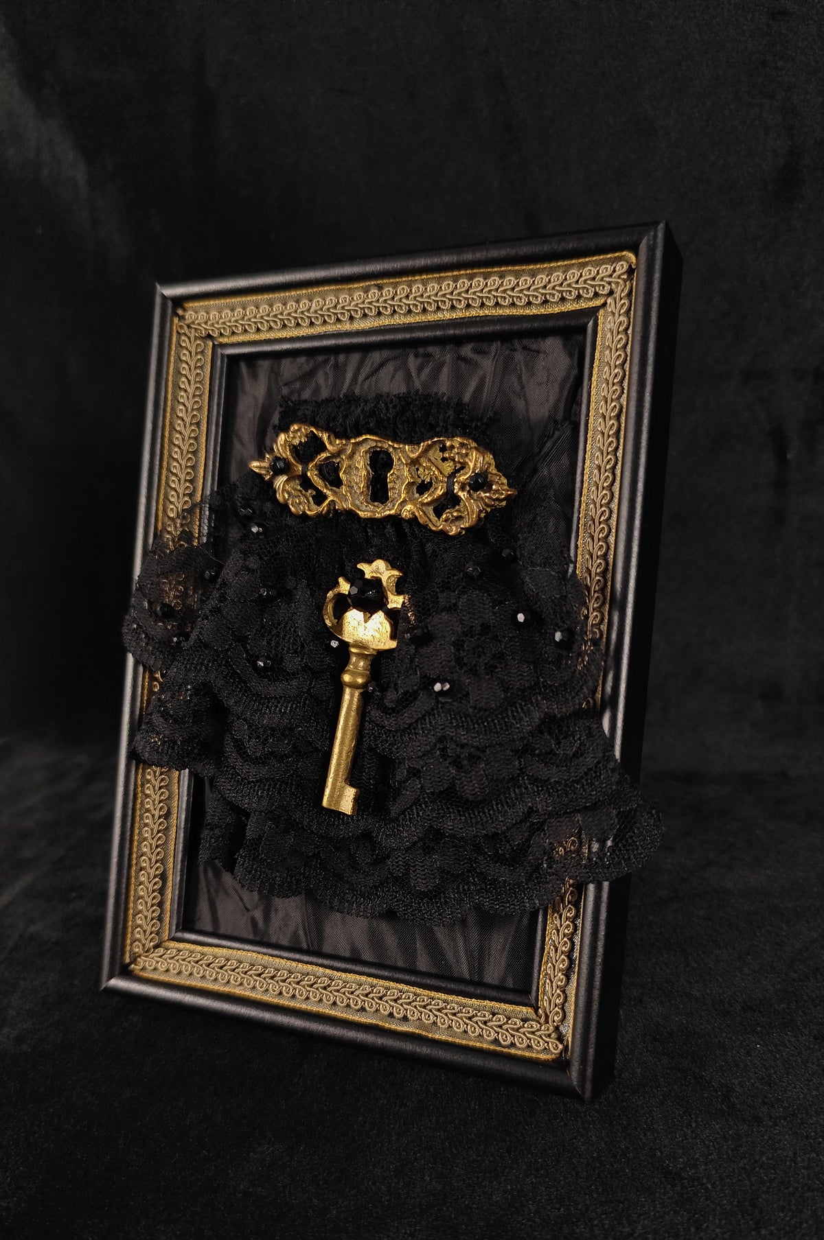 A framed gothic decorative piece featuring a black lace ruffle arrangement, a gold ornate key, mounted inside a black frame with an intricate gold border, set against a dark textured background.