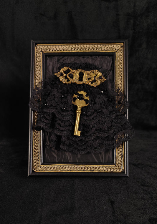 A gothic-themed frame featuring a black lace ruffle arrangement, a vintage gold key, and a gold keyhole ornament, all set against a black fabric background.