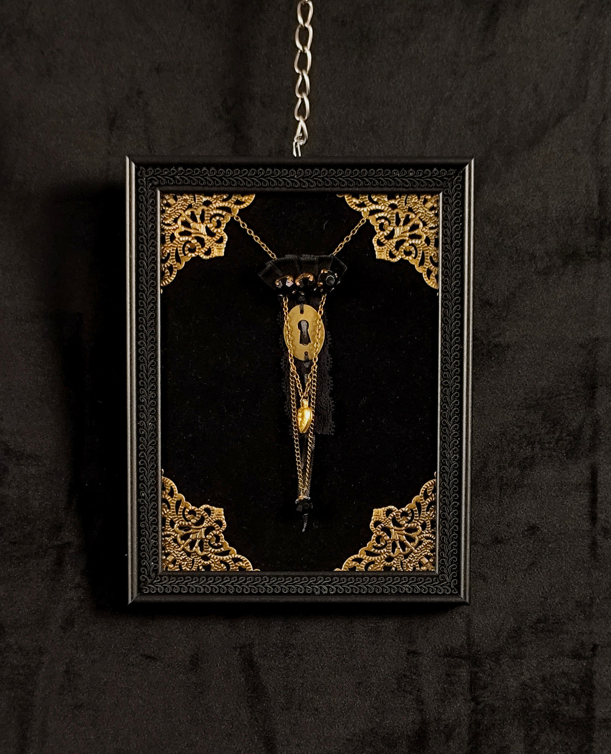 A gothic-style framed brooch displayed on a black velvet backing with ornate gold corner details, hanging against a dark textured background.