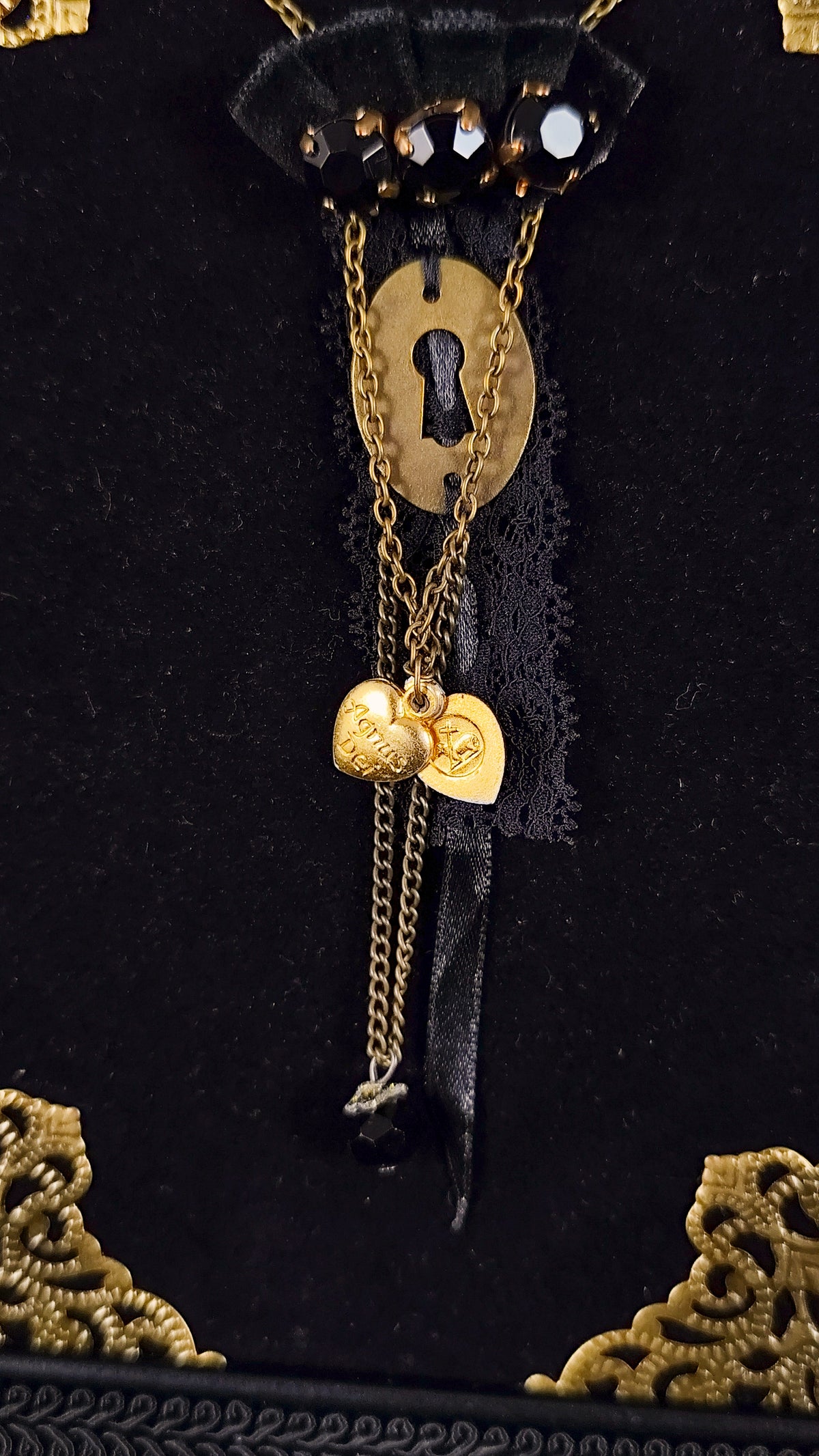 Close-up of a gothic brooch featuring a golden keyhole, agnus dei heart, chains, lace, velvet ornaments and black stones.