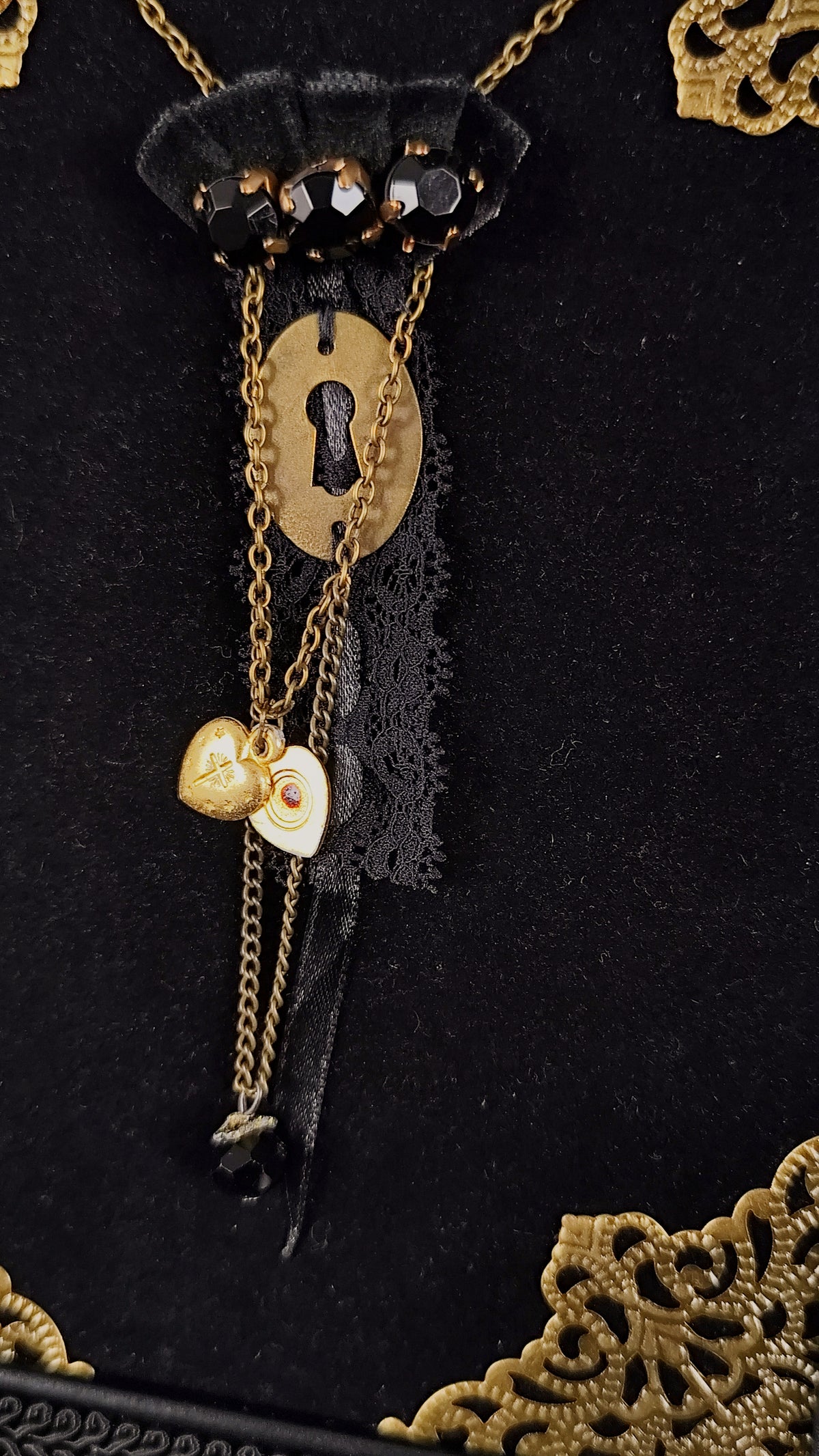 Close- up of a gothic brooch at the center featuring a golden keyhole, agnus dei heart, chains, lace, velvet ornaments and black stones.