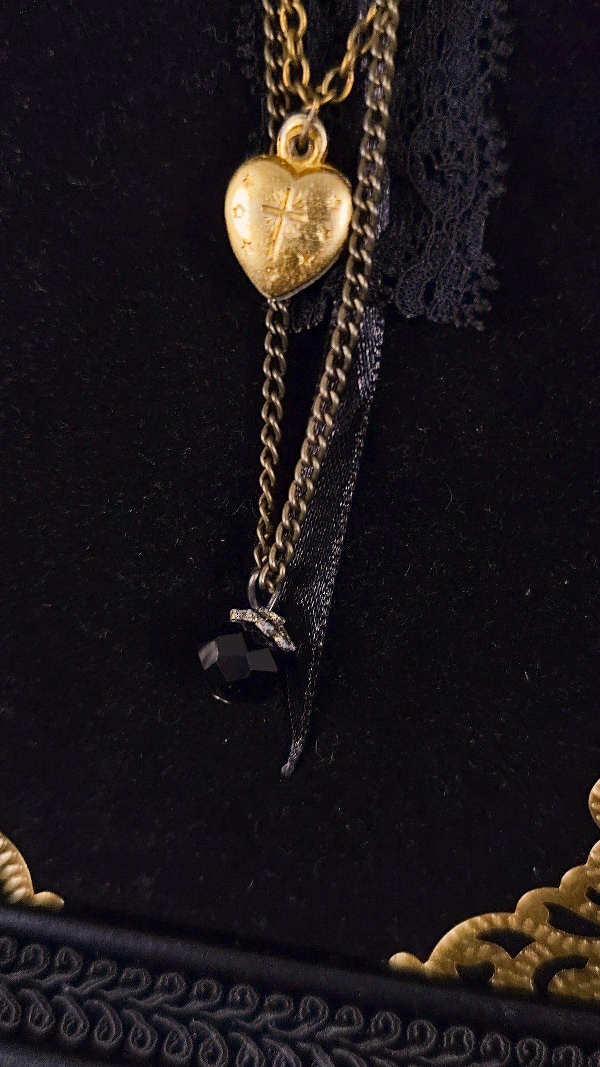 Close-up of agnus dei heart layered with a black chain and a dangling faceted black gemstone, displayed on a black velvet background.