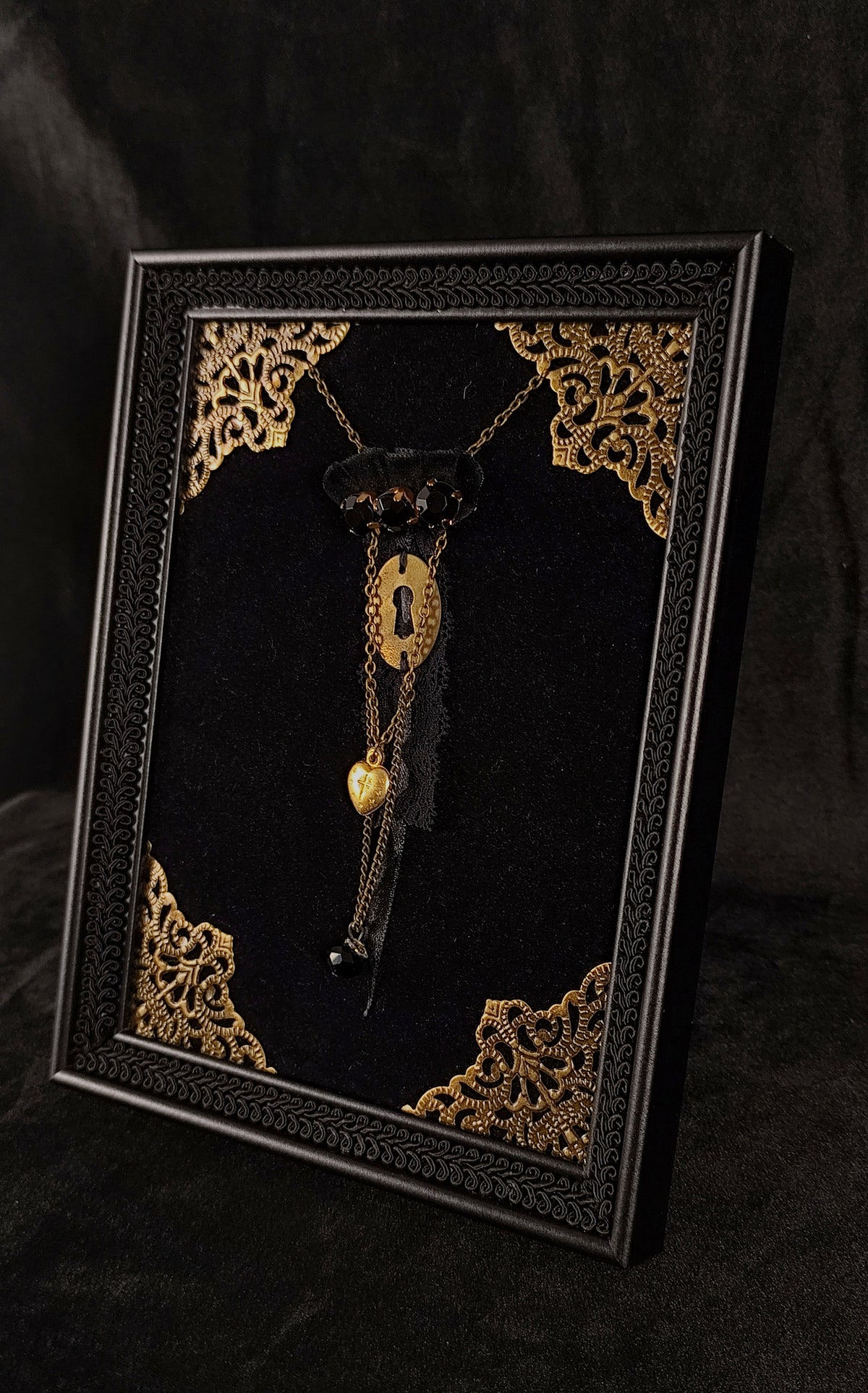 Gothic table frame with black braid, golden ornaments, a gothic brooch at the center featuring a golden keyhole, agnus dei heart, chains, lace, velvet ornaments and black stones.