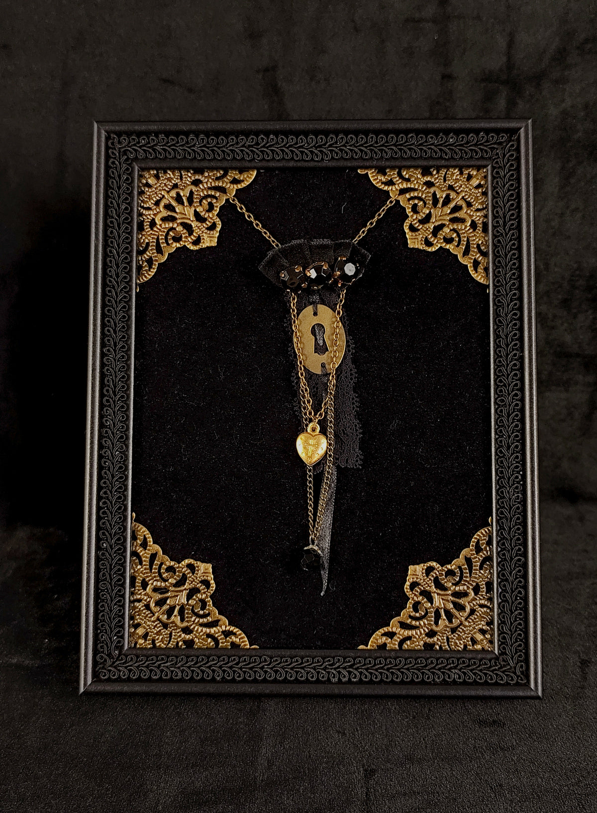 Gothic table frame with black braid, golden ornaments, a gothic brooch at the center featuring a golden keyhole, agnus dei heart, chains, lace, velvet ornaments and black stones.