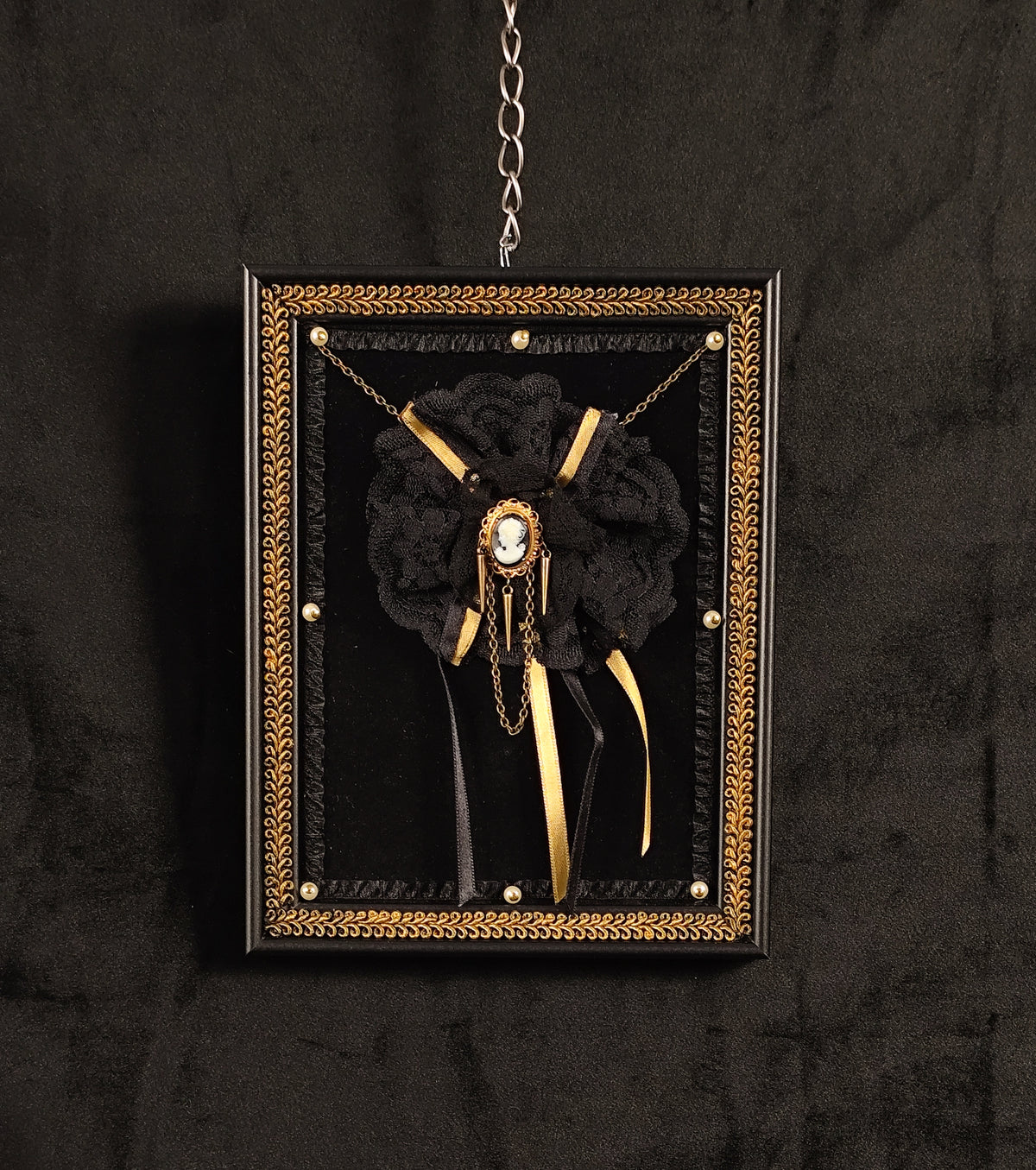 A gothic-style black floral brooch with gold chains, ribbons, and a central cameo, displayed inside a black and gold wall frame with an ornate gold border, hanging against a dark textured wall.